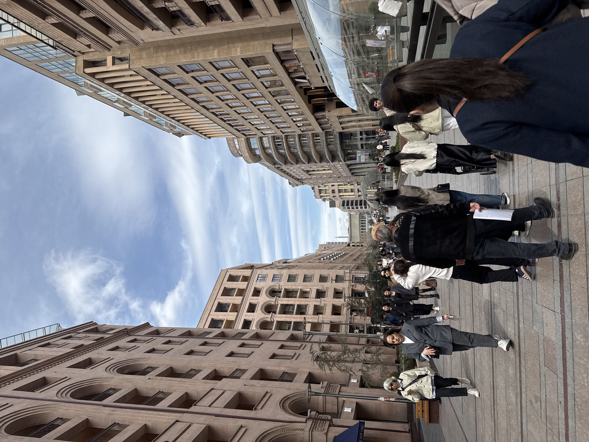 The image depicts a street scene in Yerevan, Armenia. Two large buildings flank the street, their architecture characterized by repetitive windows and arched details. A group of people is present, some standing and others walking. The sky above is partly cloudy. The buildings suggest a dense urban environment.