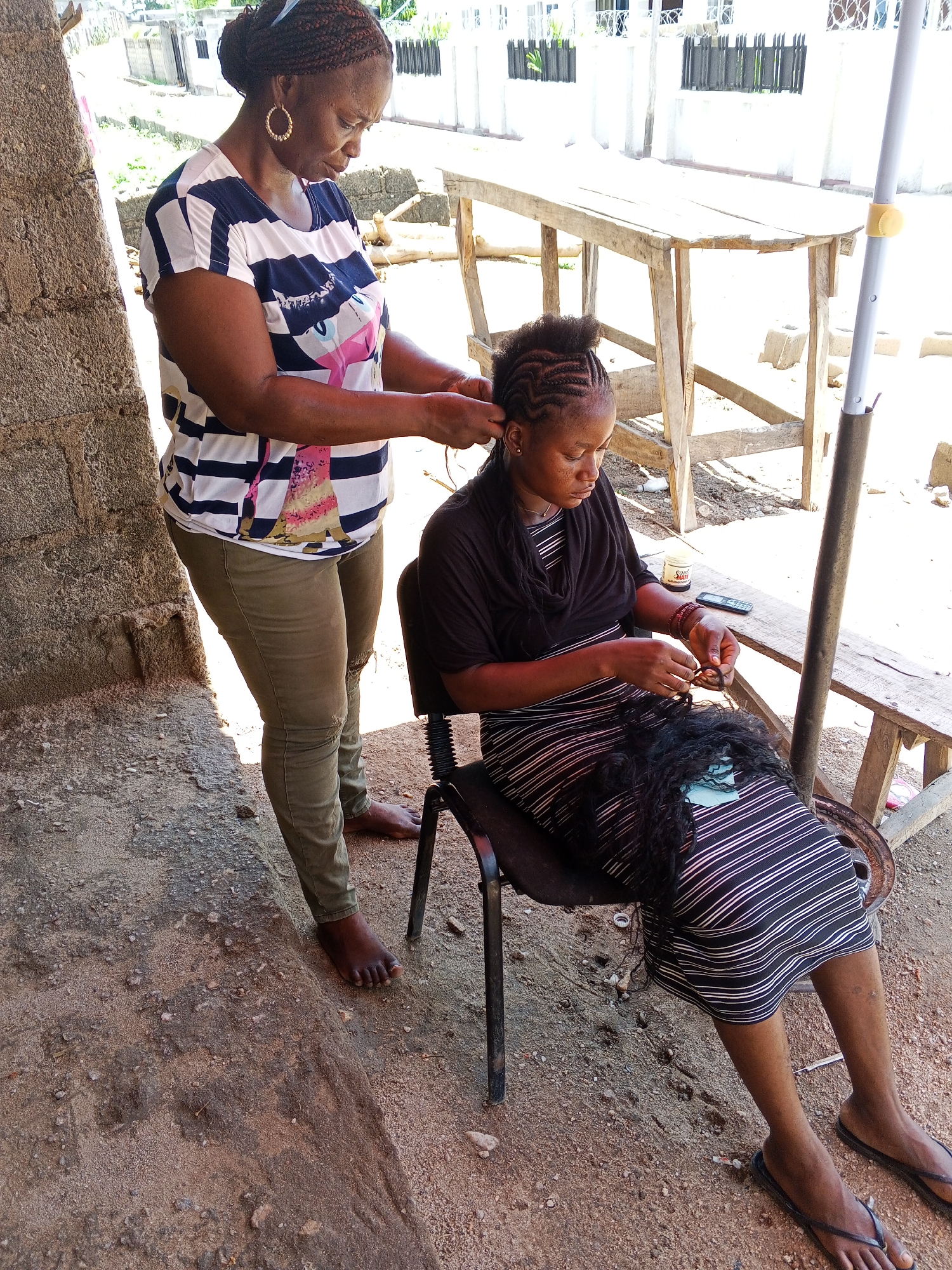The image depicts two women in Zango-ndaji, Nigeria. One woman stands while braiding the hair of the other woman, who is seated on a black chair. The standing woman wears a striped t-shirt, olive green pants, and gold hoop earrings. Her hair is styled in braids. The seated woman wears a striped dress with a black shawl. In her lap, she holds black braiding hair and appears to be working on it. A small wooden table sits to her right, holding a cell phone and a small container. The ground is covered in sand and dirt, and a concrete structure is visible in the background. Sunlight is present.