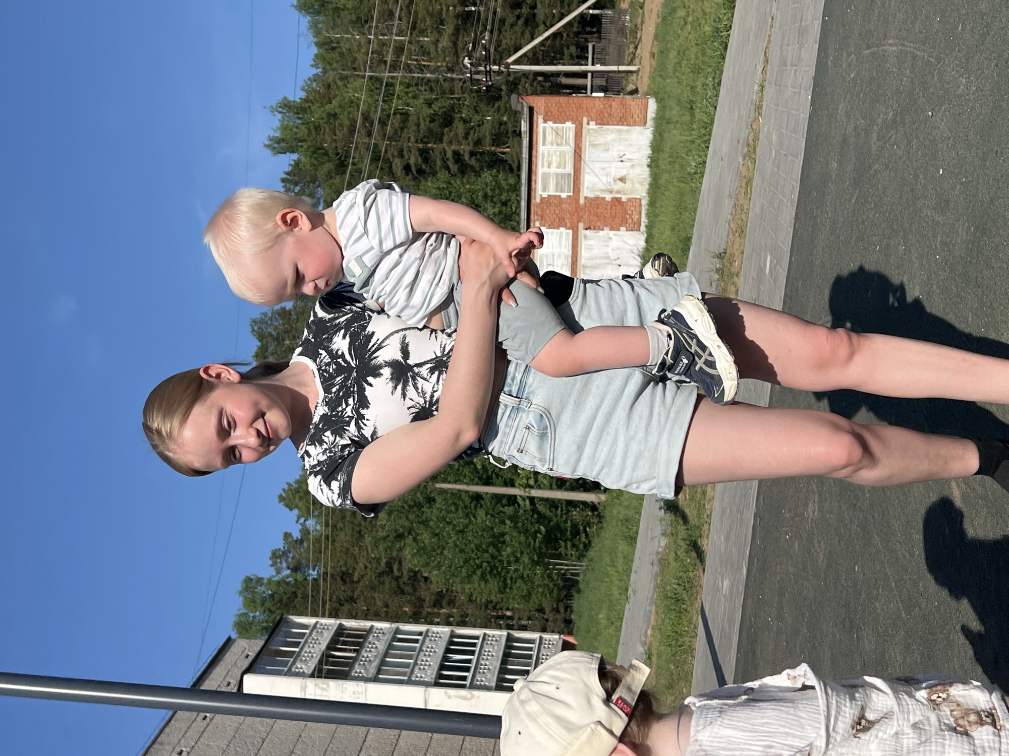 A woman is holding a young child in an outdoor setting in Noviy, Russia. The woman has light hair pulled back and is wearing a palm tree patterned shirt and denim shorts. The child has blond hair and is wearing a striped shirt and gray shorts. The background includes a building with white and red brick, trees, and a clear blue sky. Another person is partially visible at the bottom of the image wearing a baseball cap and light-colored striped shirt. The surface is paved with gray bricks and asphalt.