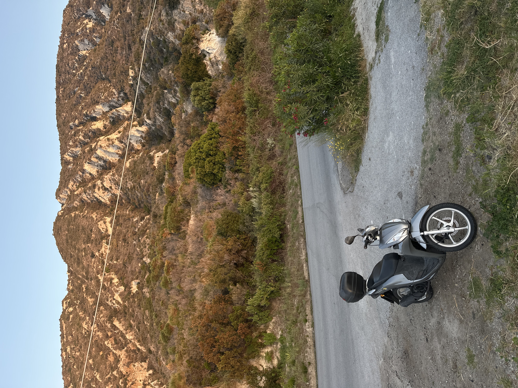 The image, when viewed in correct orientation, captures a serene mountainous landscape, likely on the island of Lipari, Italy. A narrow, two-lane asphalt road winds through the scene, bordered on its right side by a dirt and gravel shoulder transitioning into grassy patches and sparse vegetation.
In the foreground, a silver-grey scooter is parked on the dirt shoulder, facing left. It features a black seat, a black storage box mounted on the rear, and silver five-spoke alloy wheels. The word "VISION" is visible on the side of the scooter.
To the left of the road, a steep, rugged mountain face dominates the left half of the frame. The mountain is extensively covered with a mix of dense evergreen and drier, brownish shrubbery and trees, interspersed with visible, light-colored rock outcrops. A thin, light-colored cable or rope stretches diagonally across the mountain face from the lower left towards the upper right.
The sky, visible on the far left, is a clear, pale blue, suggesting fair weather. The warm, soft light illuminating the mountain and parts of the vegetation indicates either early morning or late afternoon ("golden hour"). No human activity is depicted; the scene is one of quiet, natural beauty with a parked vehicle. Along the roadside, various shrubs with small red and yellow flowers add touches of color.
