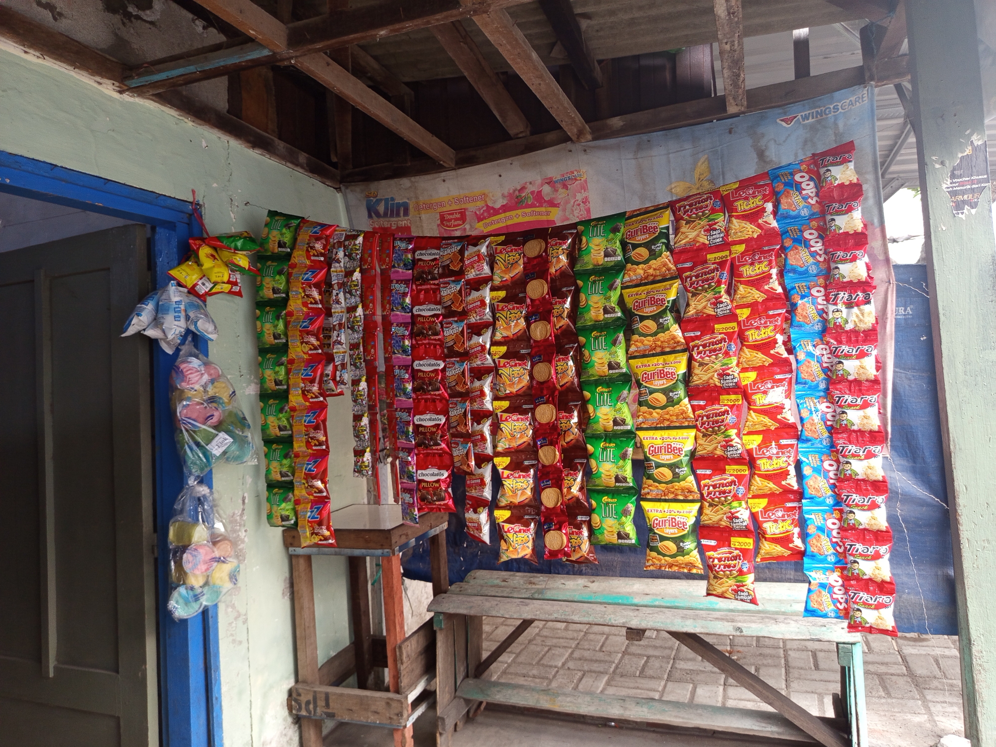 The image depicts a roadside stall in Tebel, Indonesia, likely a small warung selling snacks. The stall is characterized by a wooden frame structure with a corrugated metal roof, partially visible. The main focus is a display of various packaged snacks, predominantly chips and candies, hanging from strings on the wall. These snacks are in colorful, individually wrapped packets. To the left, a blue doorway is visible. Two wooden tables or stands are placed in front of the snack display. The ground appears to be paved with rectangular bricks. The overall impression is of a humble, open-air retail setup.
