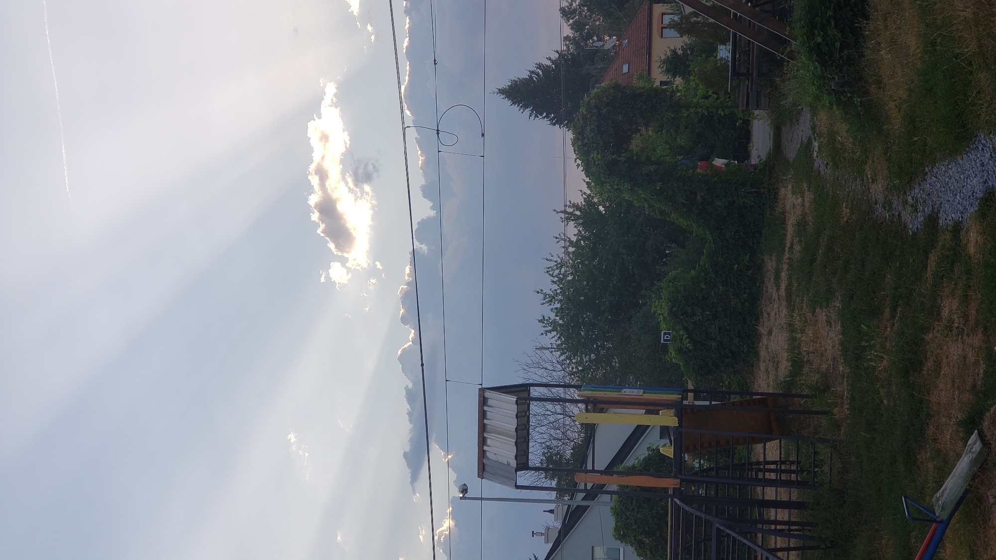 The image depicts a sky with crepuscular rays emanating from behind a cloud. Below the sky, there are several power lines strung across the frame. In the foreground, there is a children's playground structure with a metal frame and a sloped roof, partially obscured by vegetation. A seesaw is visible to the left. Behind the playground, there are trees and houses. The overall scene suggests an outdoor environment, possibly in a residential area or park. The location is identified as Prešov, Slovakia.