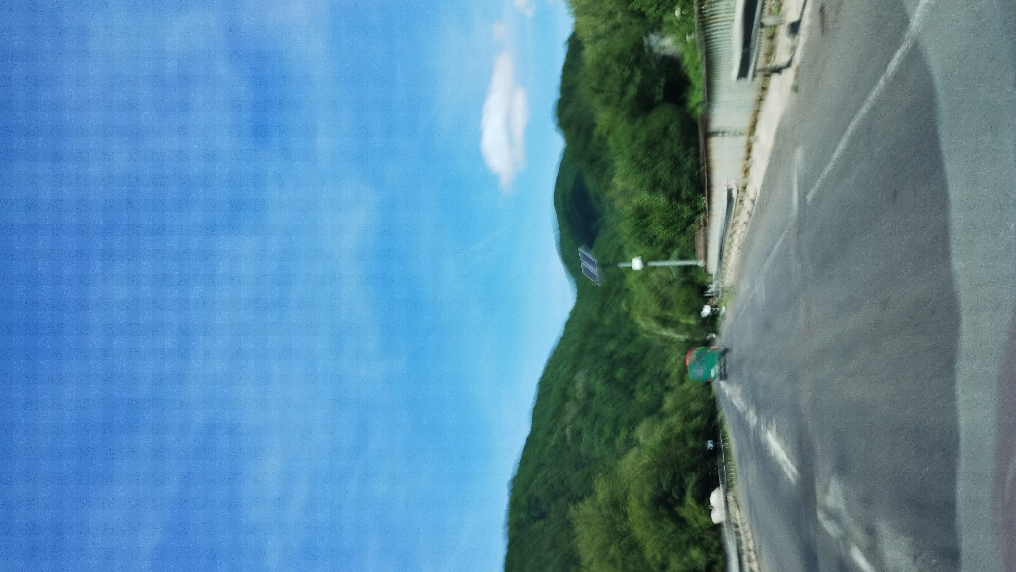 The image depicts a road in the city of Han'kovytsya, Ukraine. The perspective is from inside a vehicle, looking forward. The road is paved with visible lane markings and some imperfections. On the right side of the road, there is a metal guardrail and a fence. In the distance, a green truck is visible on the road. To the right of the road, a pole with a solar panel is mounted. Beyond the road and guardrail, a lush green forested hillside rises, with a bright blue sky with scattered clouds above. The overall scene suggests a rural or semi-rural setting.