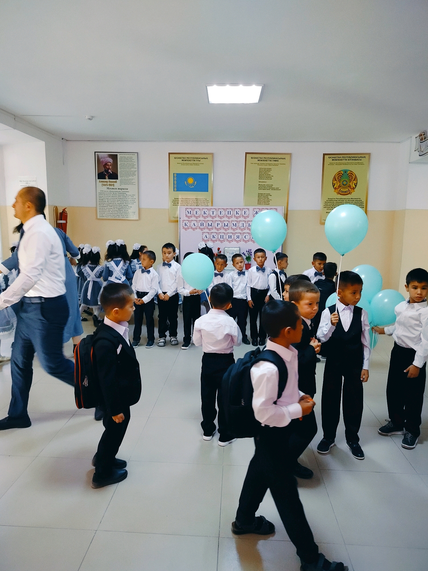 The image depicts a hallway inside a school in Kölkent, Kazakhstan. Several young boys, dressed in white shirts, black vests, and black pants, are gathered, some holding light blue balloons. A man in a white shirt and dark pants is walking away from the camera, while other students, also in school uniforms, are visible in the background. Posters are displayed on the wall, including one with a portrait and text, and another featuring the flag of Kazakhstan. The floor is tiled with light-colored square tiles. The overall atmosphere suggests a school event or the beginning of the school year.