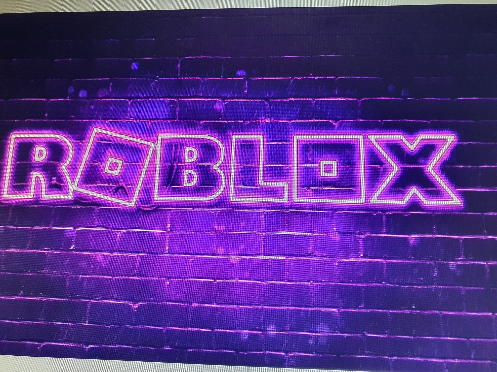 The image displays the Roblox logo rendered in neon lights against a dark brick wall. The neon lights are predominantly purple and pink, casting a vibrant glow. The brick wall appears to be a dark, textured surface, with visible mortar lines. The Roblox logo is clearly legible, with its iconic blocky lettering and characteristic square within a square symbol. There are no people or animals present in the image. The overall aesthetic suggests a modern, urban setting, possibly a storefront or an entertainment venue. The specific location in Tashkent, Uzbekistan, cannot be determined from the visual information provided in the image.