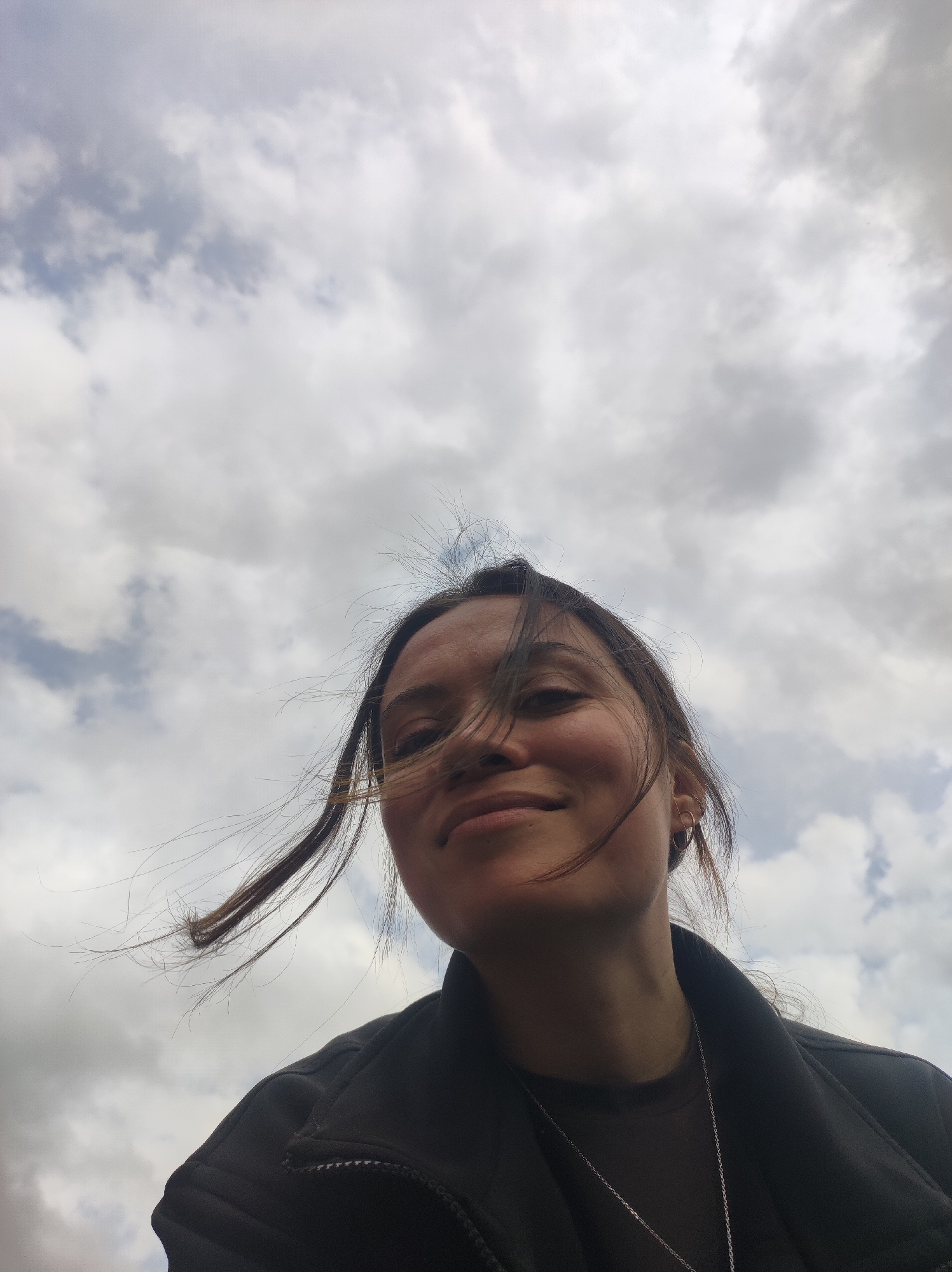 The image is a low-angle portrait of a person, likely a woman, looking up at the camera. Her face and upper body fill the lower half of the frame, with a cloudy sky dominating the background.
She has dark hair, some strands of which are visibly blown across her face and away from her head, suggesting a breezy outdoor environment. She is wearing a dark, possibly black or dark grey, jacket with a zipper and a collar over a dark top. A thin silver chain necklace with a small pendant is visible around her neck, and she has small hoop earrings in both ears.
She has a faint, pleasant smile on her face, and her eyes are directed towards the camera. The lighting creates some shadows on the left side of her face (viewer's left), while the right side is brighter, hinting at an overcast daytime setting.
The sky is filled with varying shades of grey and white clouds, indicating an overcast or partly cloudy day. The scene is outdoors. No text is visible in the image. The location context provided is Фоки, Russia.
