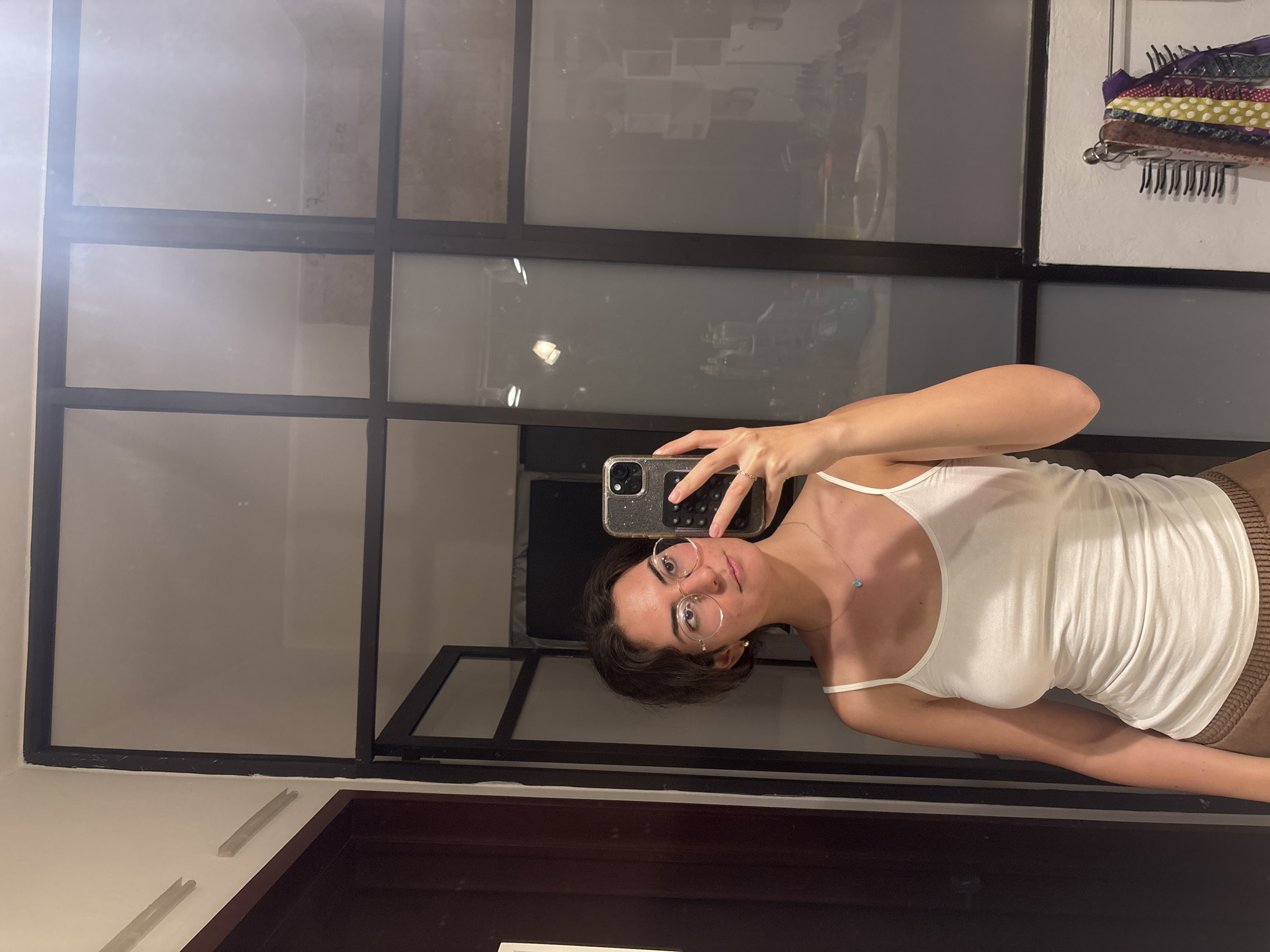 A person with short, dark hair and wearing round glasses is holding a smartphone in their right hand, taking a selfie in front of a large mirror. The person is wearing a white tank top and a necklace with a blue pendant. The mirror is sectioned with black frames, and reflections of a room with various objects and furnishings are visible. In the upper right corner, a rack holding scarves or other cloth items is attached to the wall. The lower portion of the image shows a dark wood surface, possibly a cabinet or counter. The location appears to be indoors, likely in a residential or hotel room. It is difficult to ascertain the exact location within Puebla, Mexico from this image.