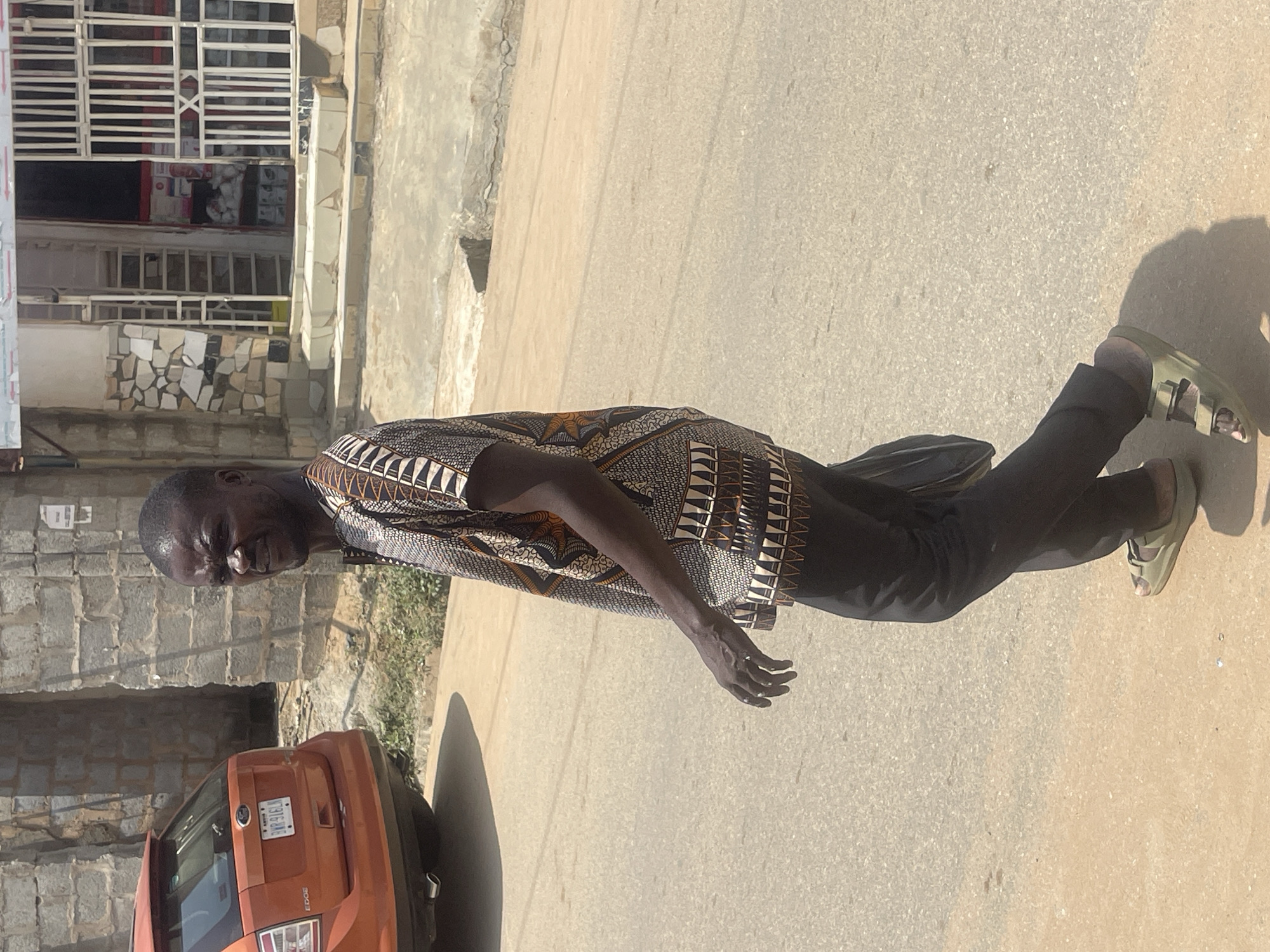 A man stands on a paved street in Abuja, Nigeria. He is wearing a patterned shirt and dark trousers, and light-colored sandals. His left arm is bent at the elbow, and his right hand is extended downwards. He is looking towards the camera with a slight smile. Behind him, an orange Ford Edge SUV is parked. To his left, a building with a white-barred facade is visible, with stone and tiled details. The scene is outdoors, and the lighting suggests it is daytime. The pavement is light brown, and there is a shadow cast from the man and the car. The overall impression is of a casual, everyday scene in an urban environment. There is text on the license plate of the car, which appears to read "TRP 916."