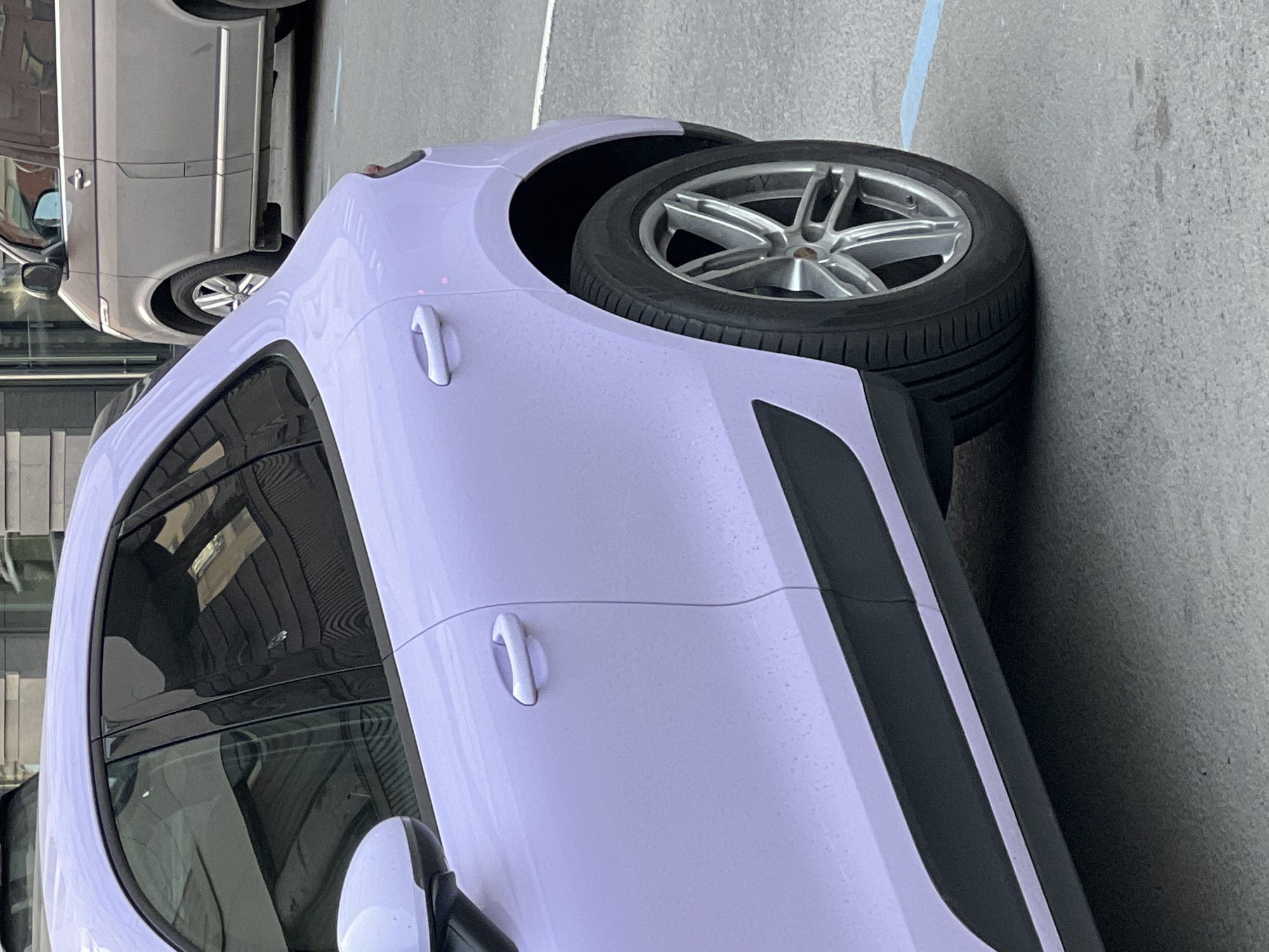 The image shows the front driver-side portion of a light purple Porsche Macan SUV parked on a paved surface, possibly concrete or asphalt. The wheel is visible and appears to have a silver alloy rim. The side of the car shows two door handles and a black accent panel towards the rear of the vehicle. Reflections are visible in the window. The edge of another vehicle, likely a silver SUV, is visible in the upper left corner. The scene is most likely in a parking area of a building in a city environment and is in the city of Saint Petersburg, Russia.