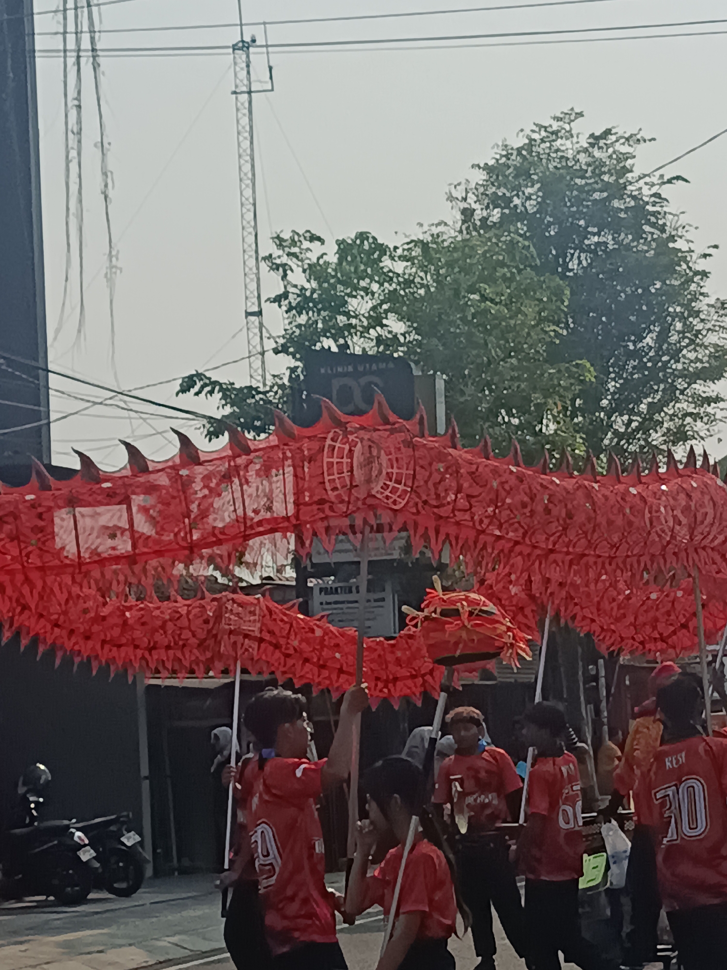 In Sleman, Indonesia, a procession is underway featuring a large, vibrant red dragon costume, carried by multiple individuals. The dragon is elaborate, with a segmented body adorned with spikes and a textured surface, possibly made of fabric and paper. Several young people, mostly male, wearing matching red t-shirts, are actively involved in maneuvering the dragon with poles. One t-shirt displays the number "30" on the back, with the word "REST" above it. In the background, a motorcycle is parked on the left side of the street, and trees with dense foliage line the right side. Overhead, power lines and a tall, lattice-like tower structure are visible against a hazy sky, suggesting an urban or semi-urban environment. Buildings with signage are also present behind the procession. The overall scene captures a community event or cultural celebration.