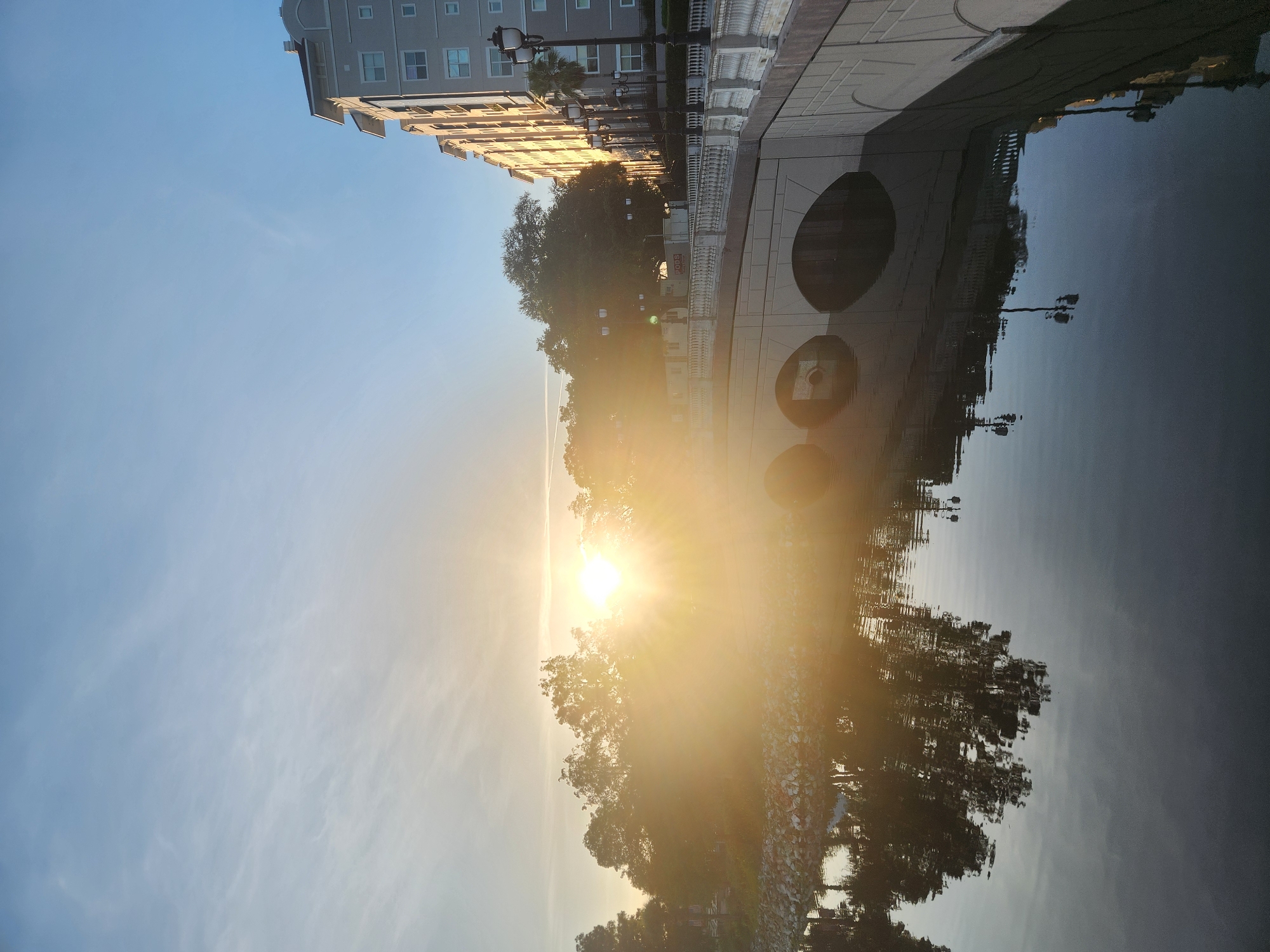 This media file depicts a serene scene, likely a waterfront area in Altamonte Springs, USA, during sunrise. The image is oriented vertically, presenting a reflection of the sky and buildings in calm water.
The subjects include a modern multi-story building with a light-colored facade and numerous windows. In front of the building, trees with lush green foliage are visible. The water acts as a mirror, reflecting these elements, as well as the sky and the bright sun.
The most prominent feature is the sun, low on the horizon, casting a warm, golden light that floods the scene and creates a sunburst effect. This indicates the time of day is early morning, likely sunrise, with clear skies and pleasant weather. The scene evokes a sense of peace and tranquility.
Notable details include decorative lampposts along the waterfront promenade and patterned paving stones. The reflection in the water is clear, showing the intricate details of the architecture and the natural elements. There is some visible text on a red sign in the reflection, which appears to read "ROSS." The overall atmosphere is one of quiet beauty and the start of a new day.