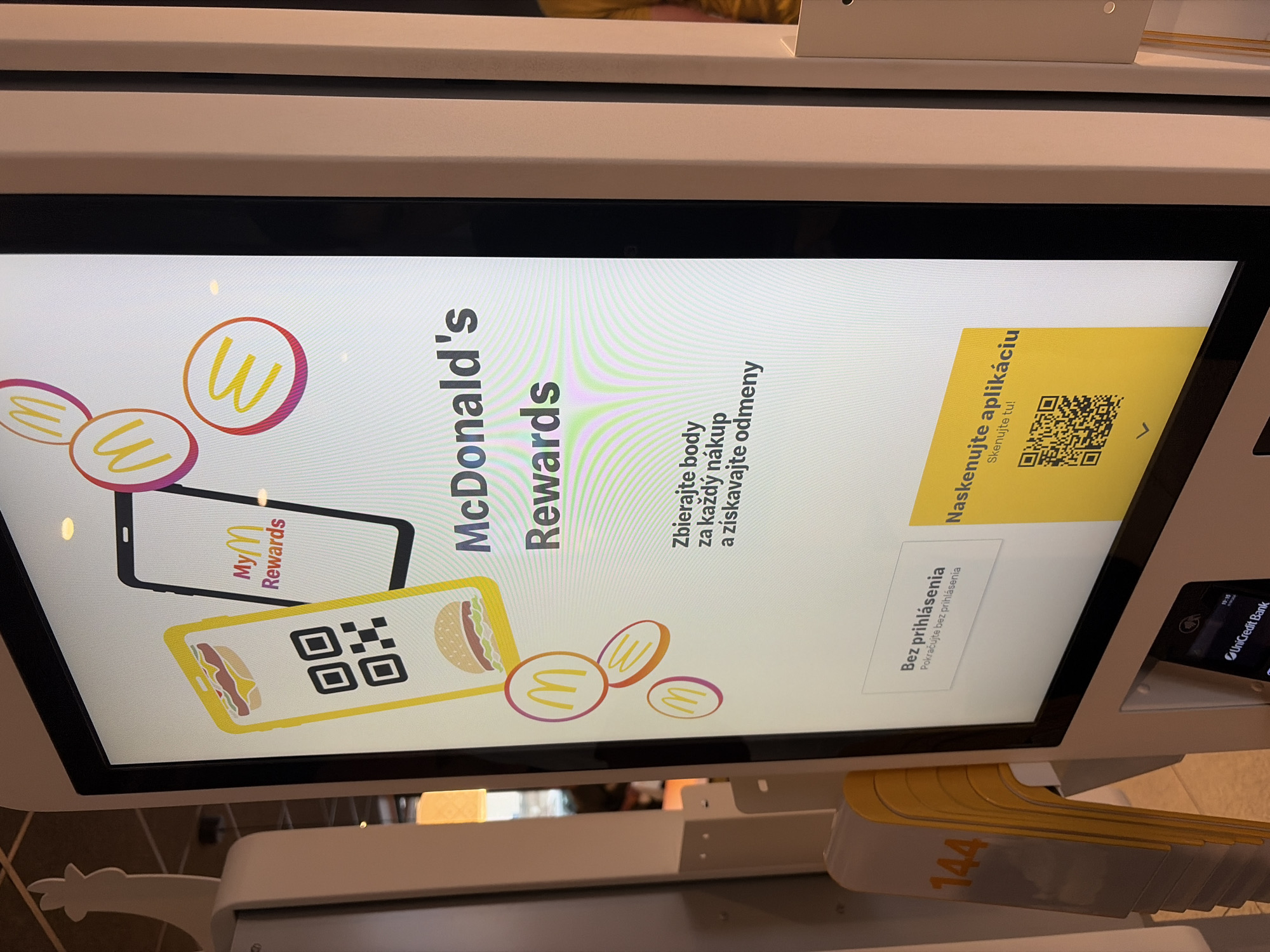 This image shows a digital kiosk inside a McDonald's restaurant. The screen displays promotional material for McDonald's Rewards, featuring a smartphone with a QR code and illustrations of burgers and McDonald's logos. Text on the screen includes "McDonald's Rewards," "Zbierajte body za každý nákup a získavajte odmeny" (Collect points for every purchase and earn rewards), and "Naskenujte aplikáciu" (Scan the app). Below the main screen, there is a card reader and a display showing "UniCredit Bank". To the left of the kiosk, yellow plastic queue holders are stacked. The overall environment suggests a modern fast-food setting. This specific McDonald's is located at Levická 2214/32, 949 01 Nitra, Slovakia. There are no people visible in the immediate foreground or interacting with the kiosk.