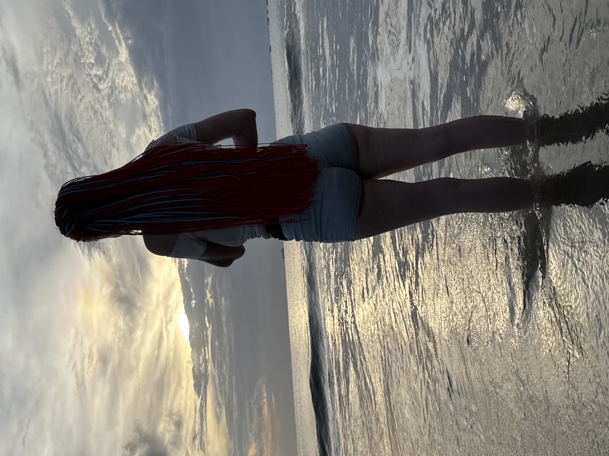 A woman stands on the beach at Kuta, Indonesia at sunset. She is positioned with her back to the camera, facing the ocean. The woman has long, red braided hair with some blue accent braids. She is wearing a light-colored top and shorts. The sky is filled with clouds illuminated by the setting sun. The ocean water is reflective and calm, with gentle waves lapping around the woman's feet.