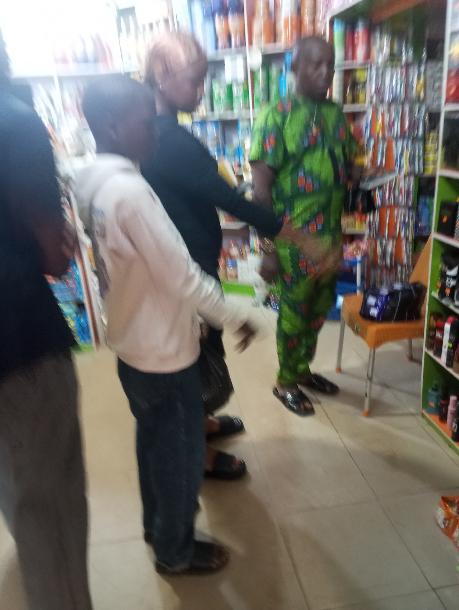 A group of people stand inside a brightly lit store. The store shelves are stocked with various products, including bottles and packaged goods. In the foreground, a person wearing a white hoodie and jeans is facing away from the camera. To their right, a person in a green patterned shirt and pants is looking at the camera. Further to the right, a woman with reddish hair wearing a black top is also present. Other individuals are partially visible on the left side of the image. The floor is tiled.