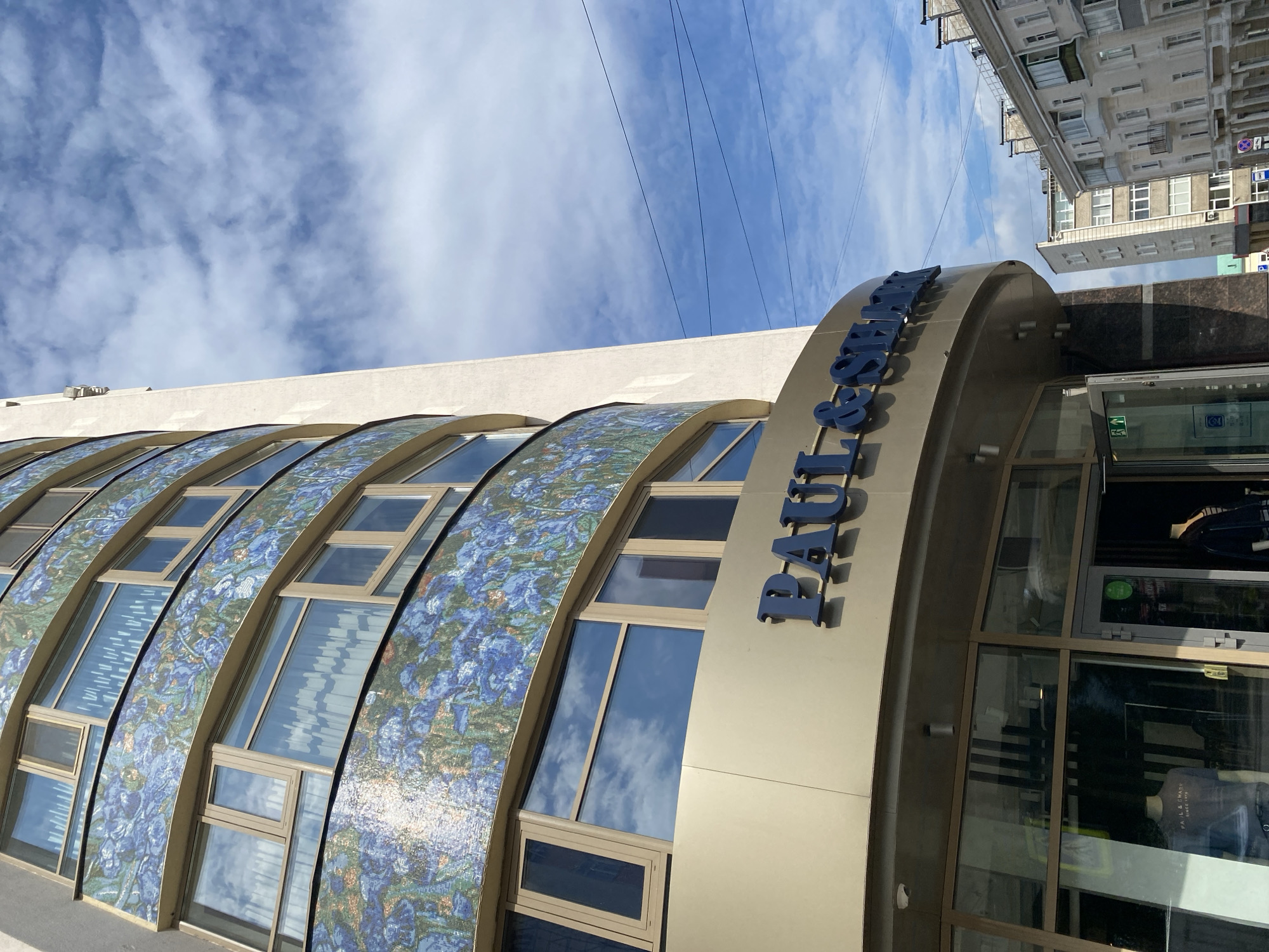 An eye-level, medium shot captures the exterior of a modern, curved building on a bright, cloudy day. The building features a beige facade with multiple floors of windows. The upper floors are adorned with decorative blue and green mosaic panels, possibly depicting flowers. Below the mosaic, a prominent beige sign with blue, three-dimensional lettering spells out "PAUL & SHARK". Beneath the sign, large glass windows reveal a store interior with mannequins dressed in casual wear. The entrance to the store is visible, with one door ajar. To the right of the main building, two older, multi-story residential buildings with balconies are visible. The sky is a vibrant blue, filled with fluffy white clouds. The scene is set in a city, likely Perm, Russia, as indicated by the signage. There are no people visible in the frame.