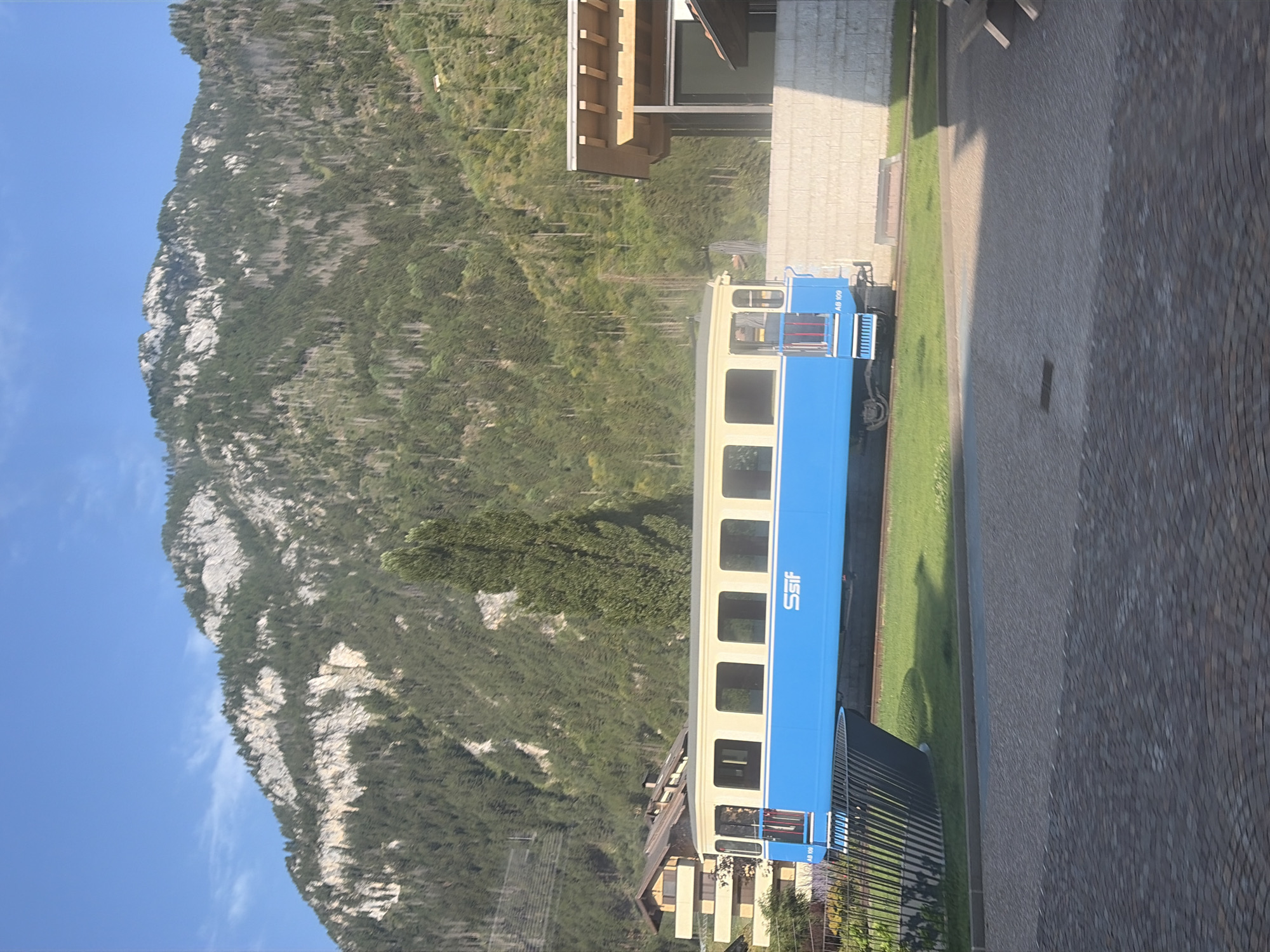 A blue and beige train car is parked on a track in Predazzo, Italy. The train car has multiple windows and the word "SSIF" is visible on its side. Behind the train, a mountain covered in trees rises against a clear blue sky. In the foreground, a paved area with a stone pattern is visible. A building with a wooden roof and a stone facade is located to the right of the train.