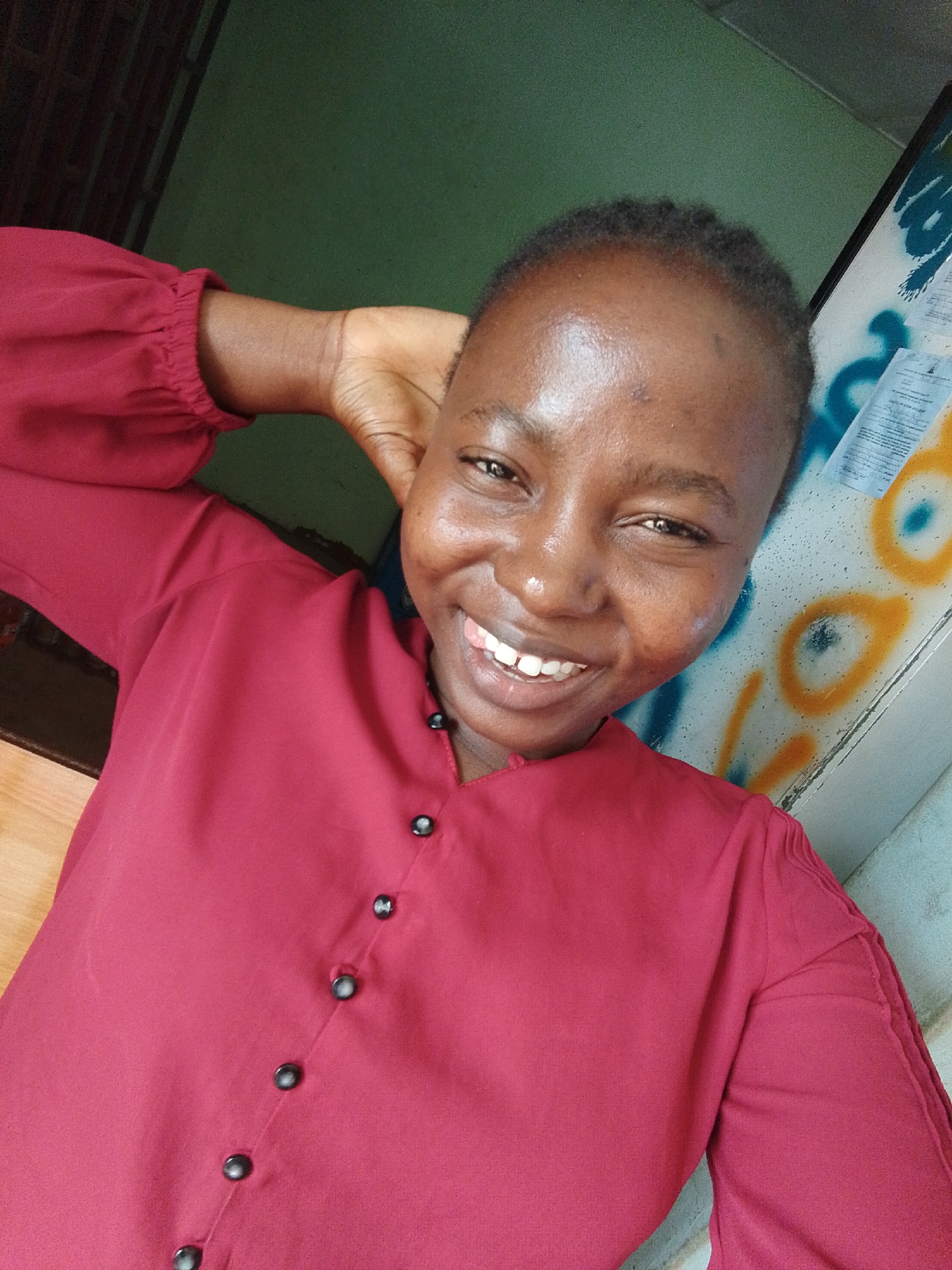 A person is smiling at the camera, wearing a red button-up shirt. Their left hand is raised behind their head. The background includes a green wall and a patterned white surface with blue and yellow markings. The location is Unknown, Unknown.
