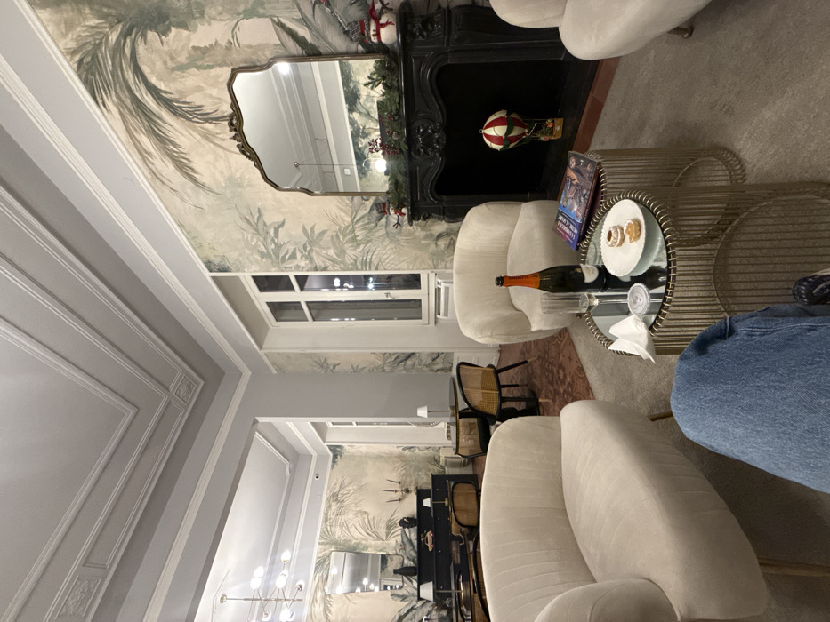 A cozy, well-lit room features cream-colored furniture, including armchairs. A round glass-topped table holds a bottle of wine, glasses, and a plate of pastries. A fireplace with a decorative mantel sits against a wall covered in patterned wallpaper. A mirror hangs above the fireplace, reflecting light and adding depth to the space. A window is visible, letting in natural light. A hallway leads to another room with similar decor.