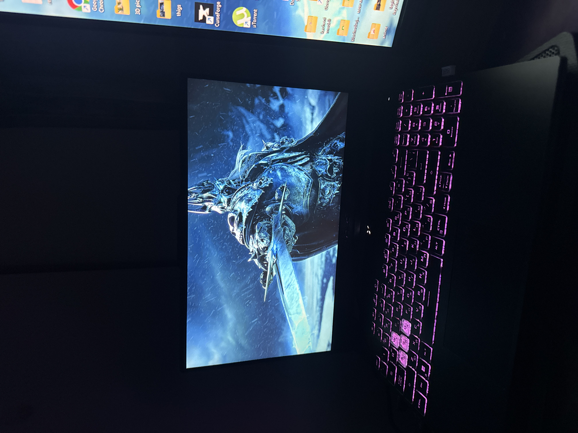 The image shows a laptop with a lit-up keyboard, and a secondary monitor or another laptop screen displaying a fantasy-style image of a figure wielding a large sword. The background is dark, and the scene seems to be indoors. The screen above the laptop screen appears to have folder and software icons on its desktop. The keyboard of the laptop is illuminated with a pink or purple light, highlighting the keys.