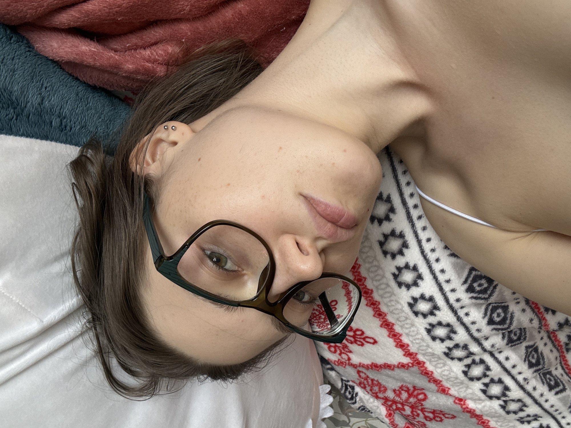 A young woman with short brown hair and glasses is lying down, looking at the camera. She has freckles on her face and two piercings in her left ear. She is wearing a white tank top. She is lying on a blanket with a red and black geometric pattern, and behind her are pillows and blankets in white, teal, and pink. The scene appears to be indoors, possibly in a bedroom. There is no identifiable information about the location within Saint Petersburg, Russia.
