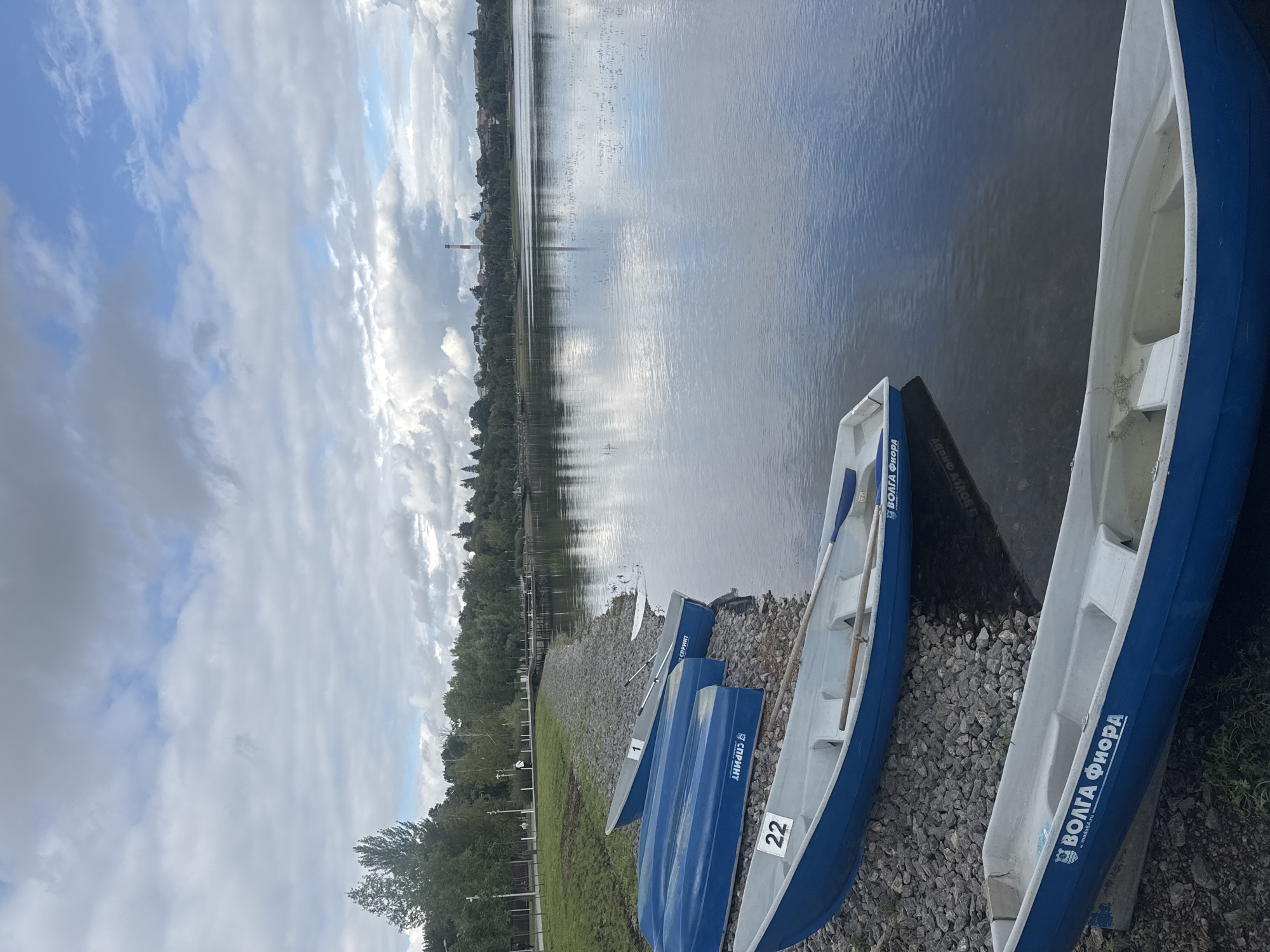 The image depicts a waterfront scene in Ufa, Russia, featuring several blue and white rowboats docked along a pebble shore. The boats are lined up in the foreground and middle ground, with some partially submerged in the water. The water itself is calm and reflects the cloudy sky above. In the background, a line of trees forms a backdrop to a distant cityscape, characterized by houses and a prominent red and white striped smokestack. A wooden pier extends into the water from the left side of the frame. The overall atmosphere is serene, suggesting a quiet day by the water.