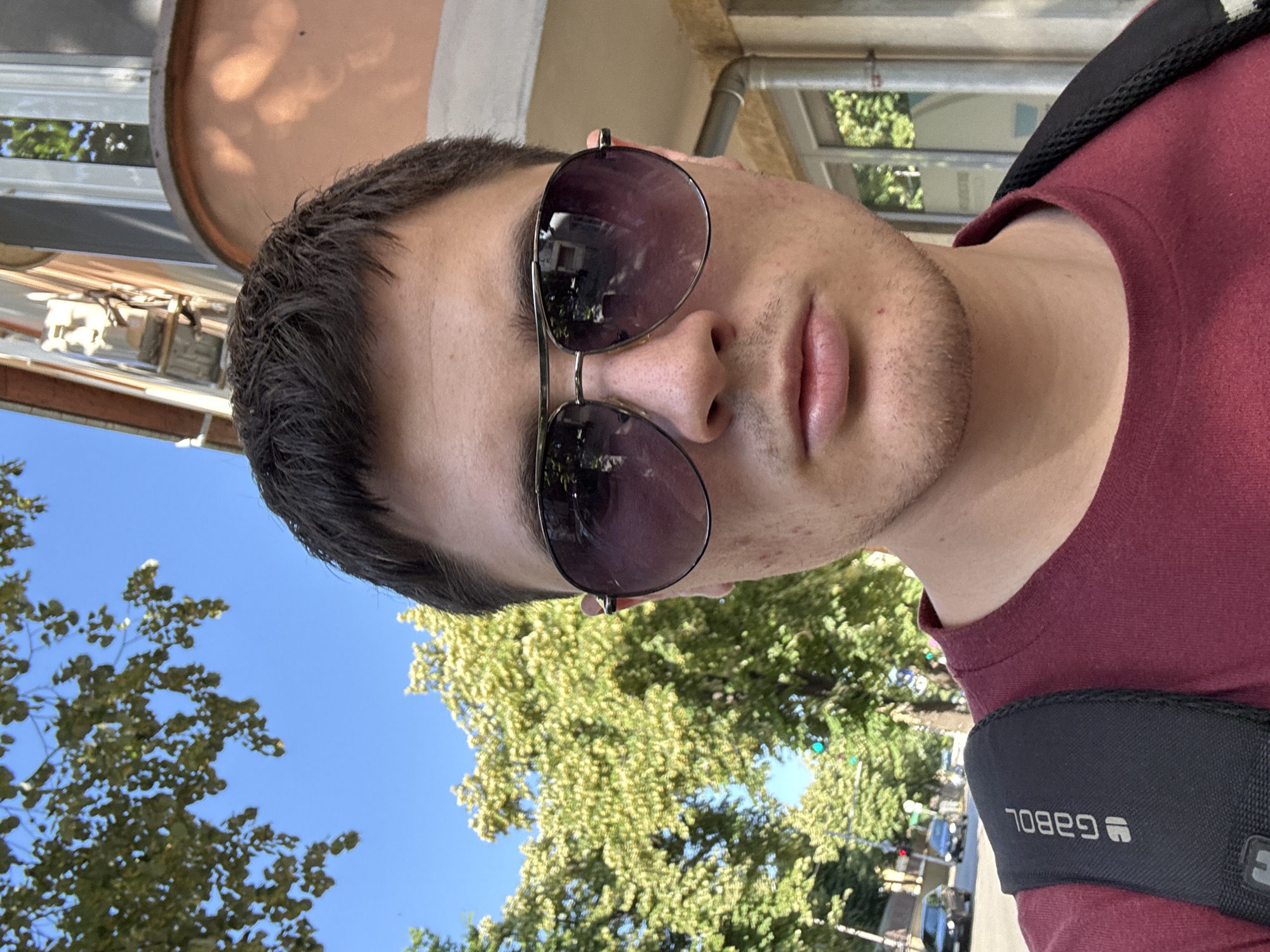 The image is a close-up, slightly angled selfie of a young man outdoors. He is wearing a dark red t-shirt and aviator sunglasses with dark lenses. His hair is dark and short, and he has light stubble on his chin. The background is a mix of blue sky and lush green trees, suggesting it is a sunny day. Behind him, hints of buildings and a street with cars can be seen, indicating an urban or suburban environment. The overall impression is casual and relaxed, possibly taken during daytime leisure. On his t-shirt, a black strap with a logo is visible, which appears to read "GABOL" in a stylized font. The image appears to be taken in Stara Zagora, Bulgaria.