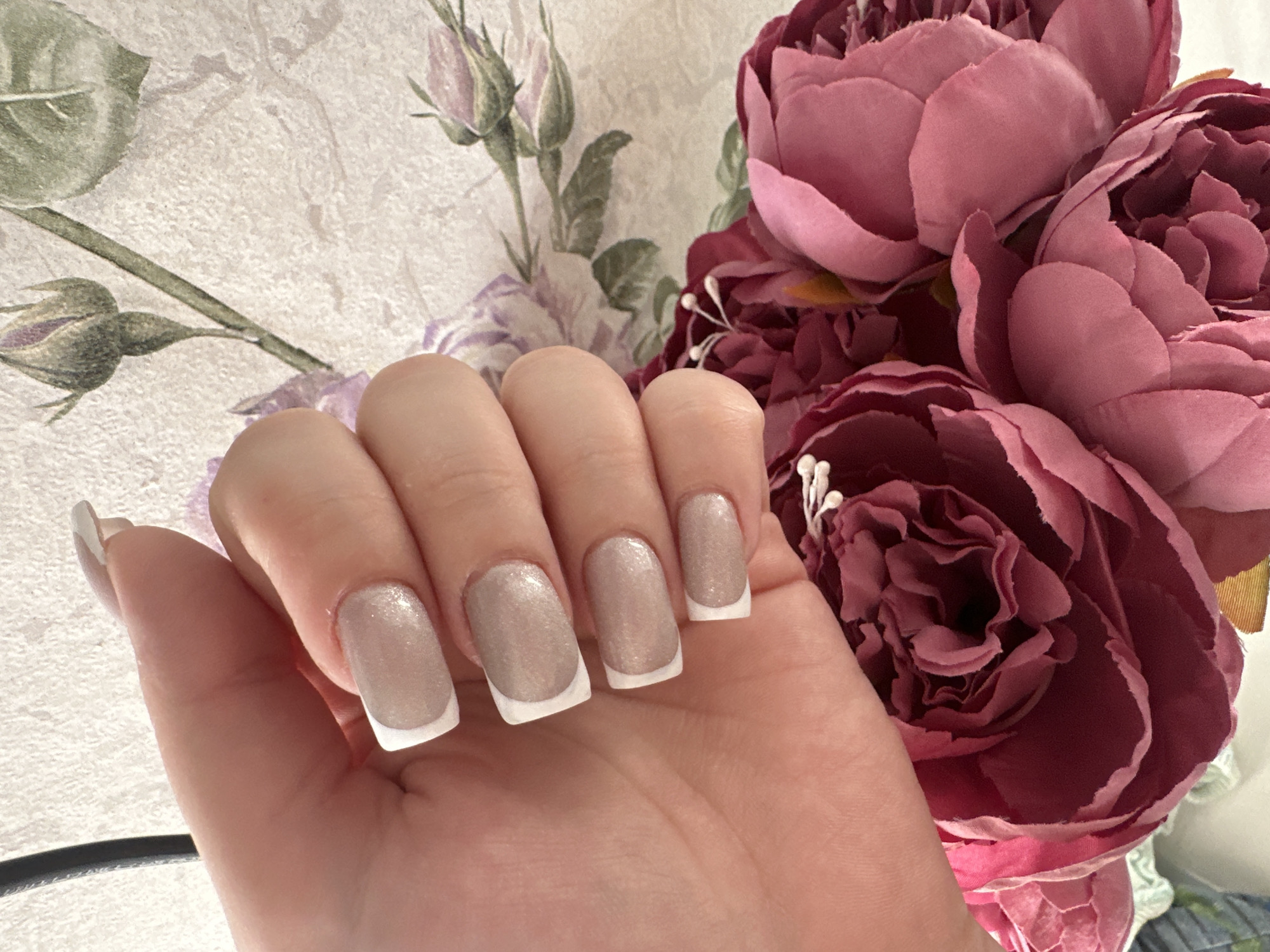 A close-up shot features a hand with manicured nails against a floral backdrop. The nails have a neutral base color with a white French tip design. The hand appears to be holding or positioned near a bouquet of large, pink flowers, possibly peonies. The background includes a patterned surface, seemingly wallpaper with a floral motif. The overall setting suggests a domestic environment. There is no indication of this image being taken in Chaikovsky, Russia.