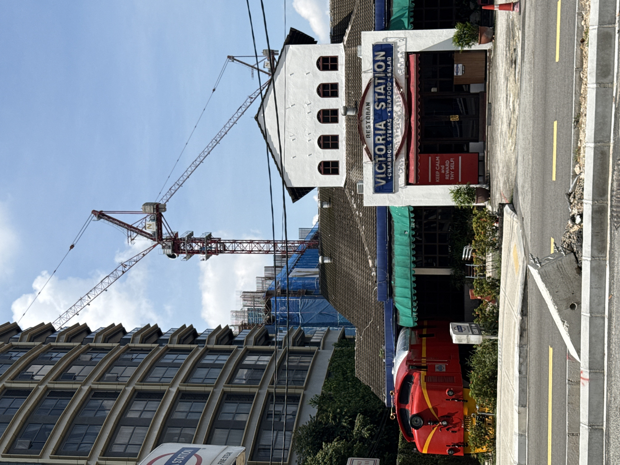 The image captures a view of the Victoria Station restaurant in Kuala Lumpur, Malaysia. The restaurant's sign is visible, and its menu includes charbroil steaks, seafood, and salad. A red locomotive train is parked near the entrance. Behind the restaurant, there's a construction site with a large crane. An adjacent building has a distinctive architectural style, featuring multiple rows of windows with protruding ledges. The sky is clear with some clouds. A road with yellow lines and zebra crossing is in the foreground.