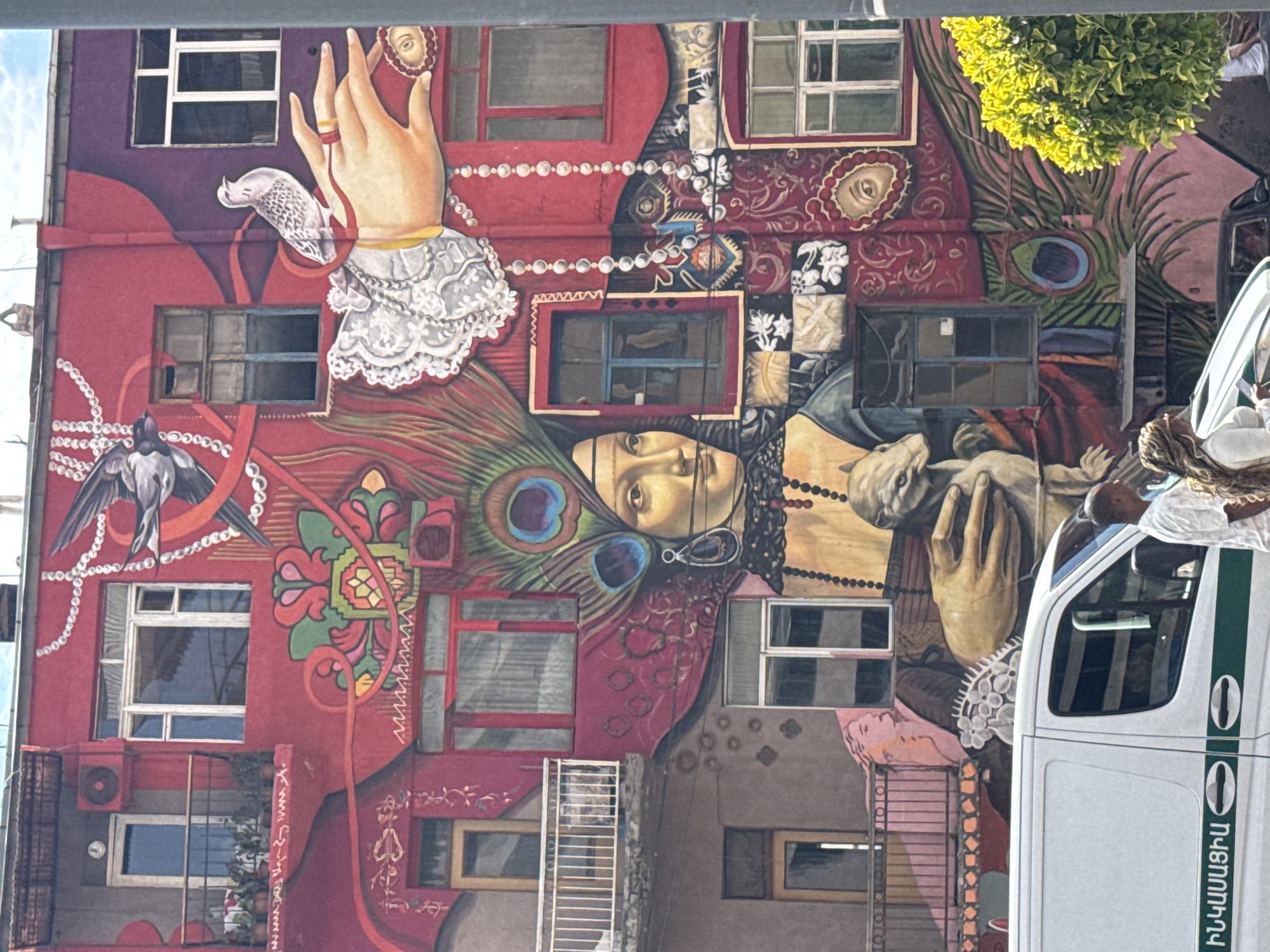 The image captures a building facade in Yerevan, Armenia, adorned with a large, vibrant mural. The mural incorporates depictions of hands, a face, peacock feathers, and bird motifs against a backdrop of windows and architectural elements. A white vehicle with green striping and Armenian text partially obscures the right side of the mural. The lighting is bright, and there's a tree visible at the top right of the frame.
