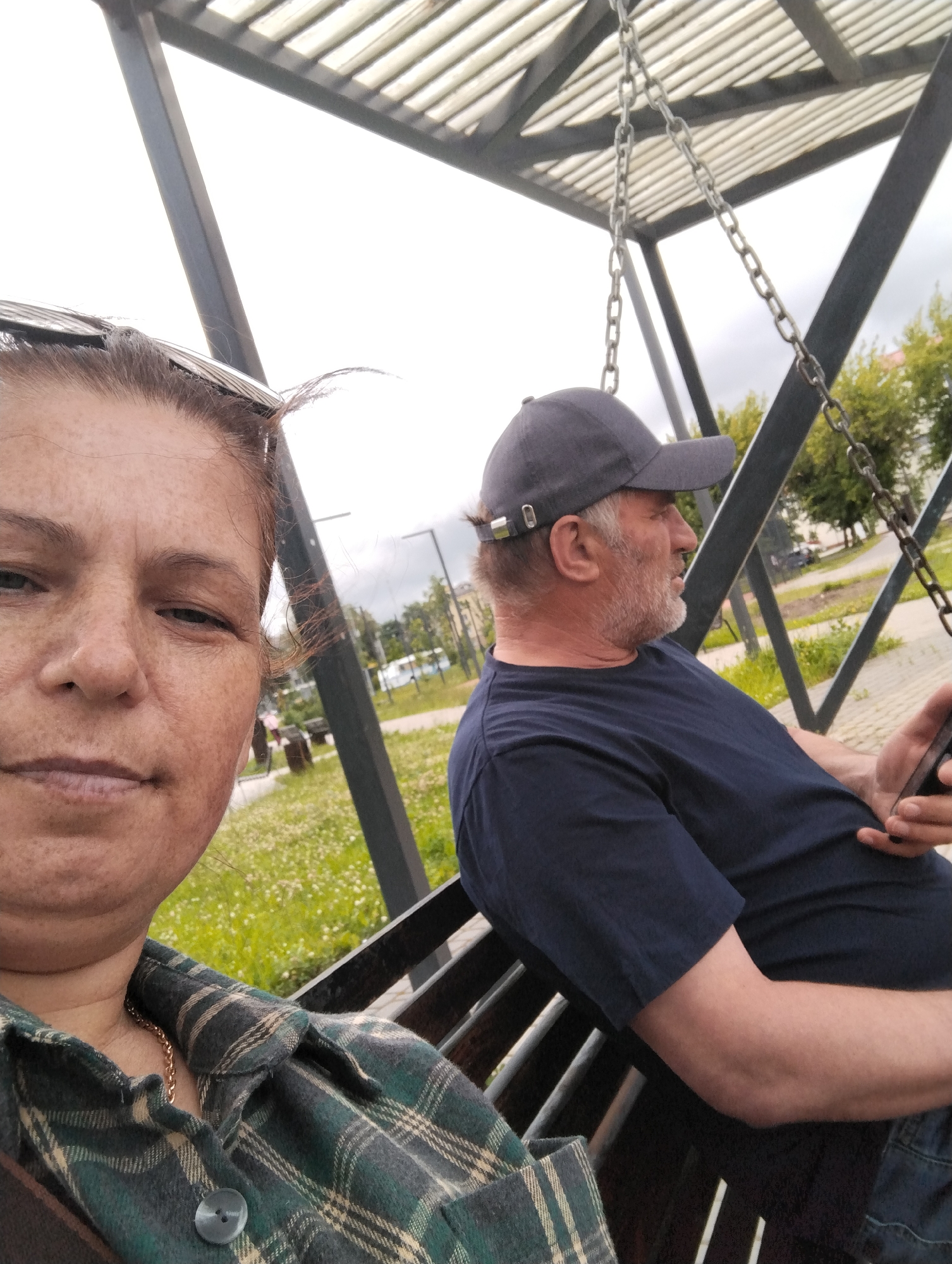 Two people are seated on a swing in an outdoor setting. On the left, a woman is close to the camera, facing it. She has brown hair and is wearing a green and white plaid shirt and a gold chain. On the right, a man is seated to the woman's right, facing away from the camera. He has a beard and is wearing a dark blue t-shirt and a grey baseball cap. He is holding a smartphone in his right hand. The swing has a metal frame and chains. The background shows a grassy area with scattered white flowers, trees, and some buildings in the distance. The sky is overcast, suggesting a cloudy day. The image appears to be a selfie taken by the woman. The overall mood seems casual and relaxed. The location is identified as Kingisepp, Russia, but no specific visual cues confirm this. There is no visible text in the image.