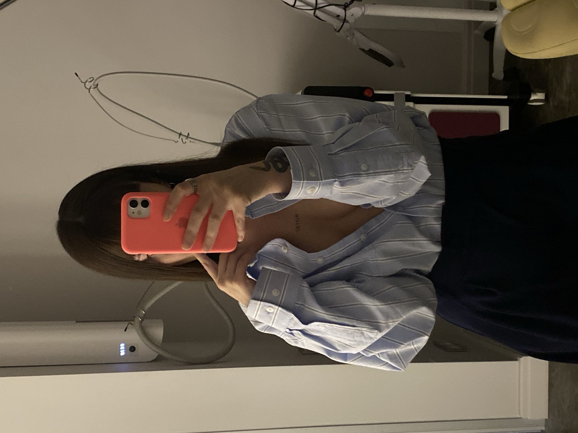 A person is captured in a mirror selfie, holding a coral-pink smartphone with dual cameras that obscures their face. They are wearing a light blue and white striped long-sleeve button-up shirt, unbuttoned to reveal a portion of their upper torso, and dark-colored lower garments, possibly a skirt or pants. Visible tattoos include a design on the inner forearm and faint numerical or textual characters, possibly "101111", on the upper chest.
The indoor setting is reflected in the mirror, featuring light-colored walls. In the upper left background, a metal hanger or part of a light fixture is visible. To the left side, below the person's arm, is a white rectangular electronic device with a blue LED display showing "00:34" and other indicators, connected by a gray tube or wire. On the right side, parts of a white-framed chair with a yellow cushioned seat are seen, along with a dark red object (possibly a cushion or component of another piece of equipment) resting on a white base. Wires are also visible in the background, suggesting specialized equipment or furniture in the room, possibly a clinic or spa.