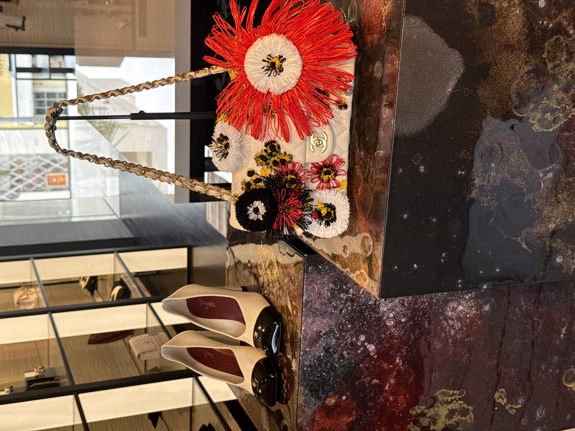 The image shows a Chanel handbag with floral embellishments on display in a Beverly Hills retail environment. The bag, primarily white with a gold chain strap, is decorated with large, colorful flowers crafted from thread and pom-poms. It sits atop a multi-toned, reflective surface, likely a display table, alongside a pair of light-colored Chanel shoes with black embellishments. A mirrored display case filled with other Chanel products is visible in the background.