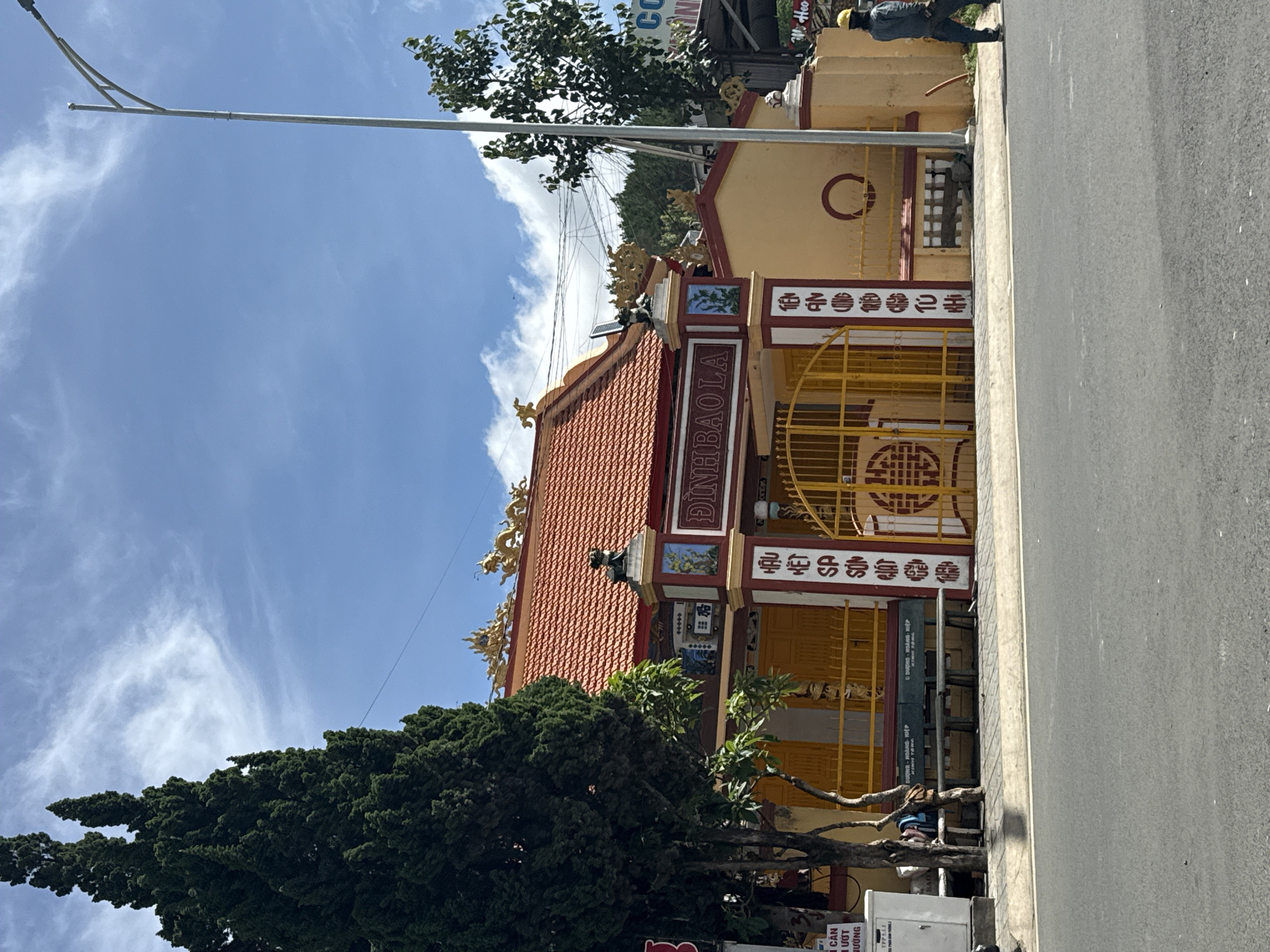 This image captures a vibrant, traditional Vietnamese temple or communal house, known as "DINHBAOLA," situated along a paved road in Da Lat, Vietnam, under a bright, sunny sky with scattered white clouds.
The central subject is a brightly colored building with a prominent orange-red tiled roof, adorned with golden dragon sculptures along its ridge. The building's walls are painted a soft yellow, complemented by red and white decorative trim and ornate pillars. The main entrance features a striking yellow metal gate, centrally embellished with a red traditional "Shou" (longevity) symbol. Flanking the gate are two large, rectangular pillars, predominantly red and white, inscribed with vertical Vietnamese characters; the left pillar bears "HẬU KẾT SƠN SƠN," while the right displays "TIÊN ẨN ỨNG CẢNH TÙ HI." Above these pillars, smaller statues, one resembling a mythical creature, sit on the roofline. To the left of the gate, multiple yellow-painted panels suggest closed doors or windows. A dark green wooden bench, inscribed with "DƯƠNG - HOÀNG HIỆP," is positioned in front of the building, to the left of the gate, next to a white utility box.
On the far left, a tall, slender evergreen tree with dense foliage reaches high into the sky, partially obscuring the building. Further to the right of the gate, another section of the yellow wall features a large, red, incomplete circular design.
In the foreground, an asphalt road stretches across the frame, marked by a white line on its edge, separating it from the paved sidewalk in front of the building. On the far right, a person in blue work attire and a yellow hard hat is seen walking away from the viewer along the sidewalk. Another individual is potentially seated on the bench on the far left, mostly hidden by the tree and utility box, with a blue backpack partially visible.
A tall, silver lamppost stands prominently on the right side of the road, extending an arm over the street. Overhead electrical wires crisscross the blue sky, particularly dense on the right side above the lamppost and a smaller, leafy tree. Distant hills covered in dense green foliage are visible beneath the horizon in the background.