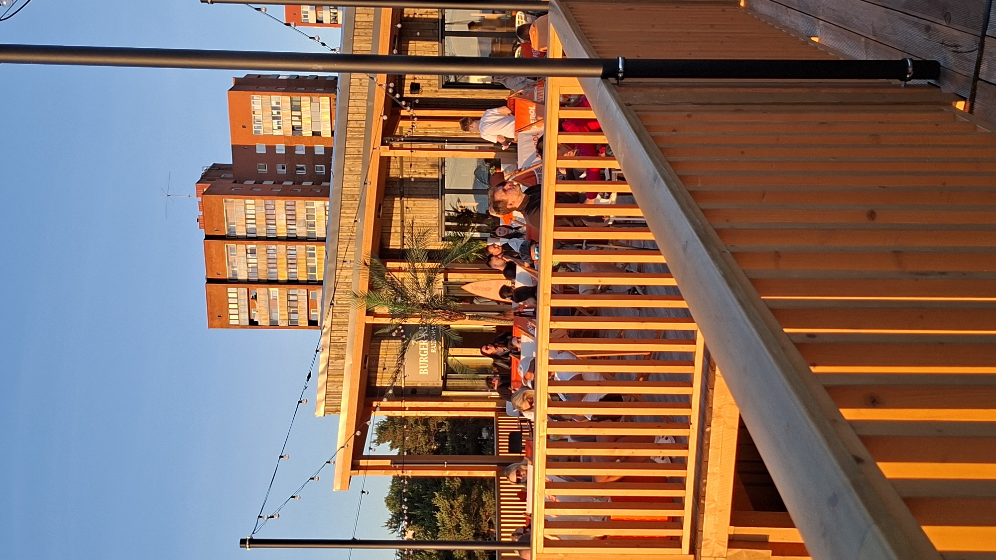 This is a vertical, eye-level, outdoor photograph taken at sunset in Tallinn, Estonia. The scene depicts a lively outdoor seating area of a restaurant or bar, possibly located near a beach or waterfront, as indicated by the casual seating and palm trees. The warm, golden light of the setting sun bathes the scene, casting long shadows and highlighting the wooden structures.
In the foreground, a light-colored wooden railing and vertical slats of the outdoor seating area are prominent, bathed in the orange glow of the sunset. Beyond the railing, people are seated at tables and on deck chairs, enjoying the evening. Some are engaged in conversation, while others appear to be relaxing or watching the surroundings. A few individuals are standing.
The mid-ground features a wooden-clad structure with large windows, likely the main building of the establishment. A sign for "BURGER K" is visible on the facade, suggesting it is a burger restaurant. Palm trees and decorative string lights add to the ambiance. A surfboard leans against a palm tree.
In the background, two multi-story, orange-brick apartment buildings are visible against a clear blue sky. These buildings appear to be of Soviet-era architecture. The setting sun casts a warm glow on their facades. Another building, possibly part of the same complex or a separate structure, is partially visible to the far right.
The overall impression is one of a pleasant, social gathering in a vibrant urban setting during a beautiful sunset.