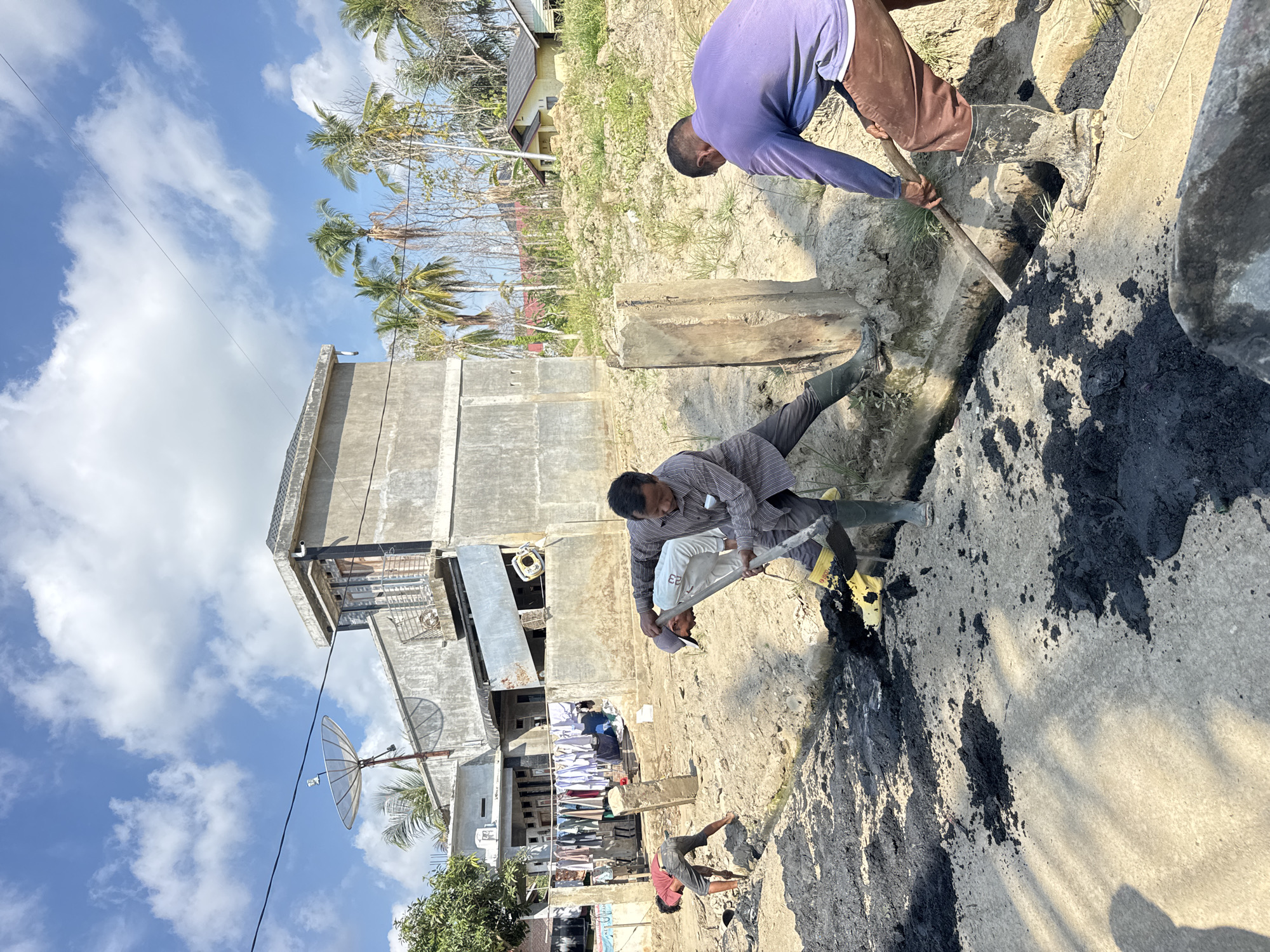 In Meunasah Raya, Indonesia, three men are working on a construction site. The ground is dry and dusty, with piles of dark soil and some scattered debris. The men are dressed in casual clothing and are wearing rubber boots, suggesting they are engaged in manual labor. One man in the foreground, wearing a purple long-sleeved shirt and brown pants, is using a shovel to move dark soil. To his left, another man in a striped long-sleeved shirt and dark pants is also using a shovel, appearing to dig into a trench. Further in the background, a third man in a red shirt and dark pants is bent over, possibly working with a tool on the ground.
The environment suggests an outdoor, semi-urban setting. In the background, there are several buildings, including a two-story concrete structure with a satellite dish on its roof, and other houses with tiled roofs. Palm trees are visible, indicating a tropical climate. Clothes are hanging on a clothesline between two structures, suggesting residential activity nearby. The sky is bright blue with scattered white clouds, and the sun is casting shadows, indicating it is daytime. The overall scene depicts manual labor on a construction project in a rural or semi-urban area of Indonesia.