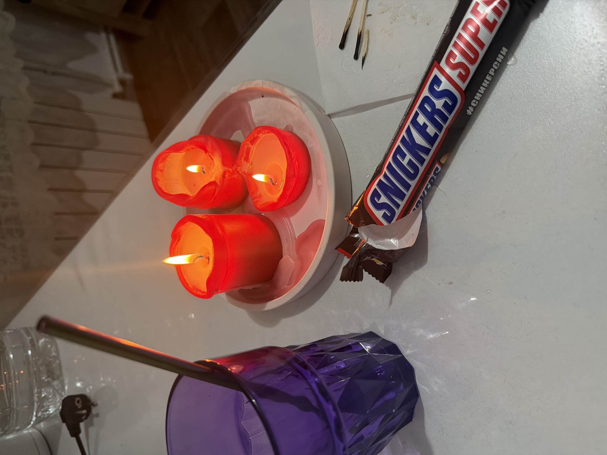 The image shows a still life arrangement on a white surface. From left to right, there's a partial view of a transparent glass, a black electrical plug with a cord, a purple faceted plastic cup with a metal straw, a white plate holding three lit red candles of different sizes, a partially unwrapped "Snickers Super" candy bar, and three spent matches. The text on the candy bar includes "#СНИКЕРСНИ" which is likely related to the Russian spelling of the Snickers brand. This suggests the location is in a Russian-speaking region. Given the text, a reasonable assumption is that the location is Leningradskaya, Russia. The scene is illuminated by the candlelight. The composition suggests a personal or intimate setting.