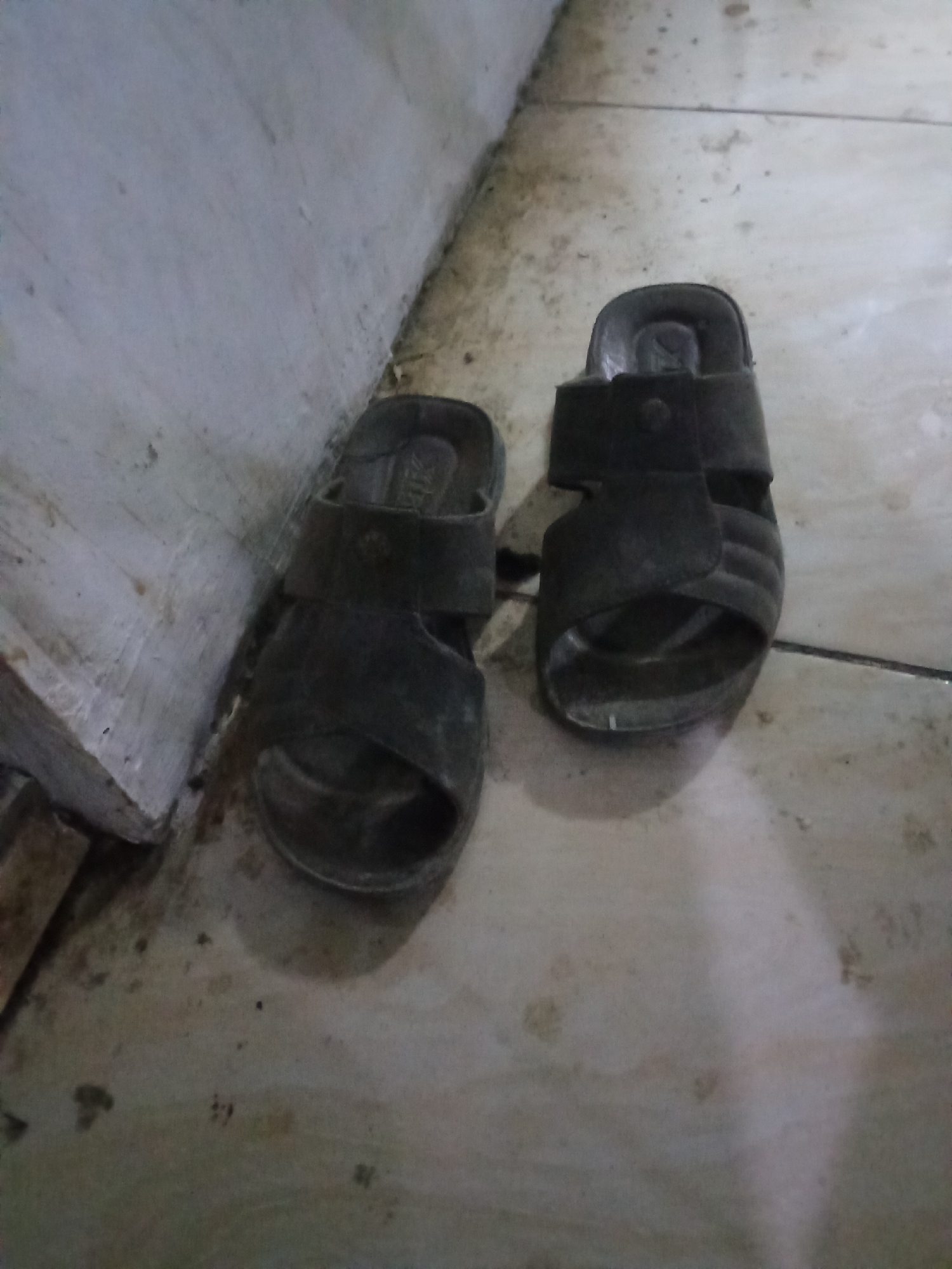 The image shows a pair of dark-colored, worn sandals on a tiled floor. The floor appears to be dirty with some smudges and debris. To the left of the sandals, there is a rough, textured wall, possibly made of concrete. The lighting is dim, suggesting an indoor environment, possibly a hallway or a porch in a residential setting. There are no people visible in the image. The scene is located in a part of the city of Pemalang, Indonesia.