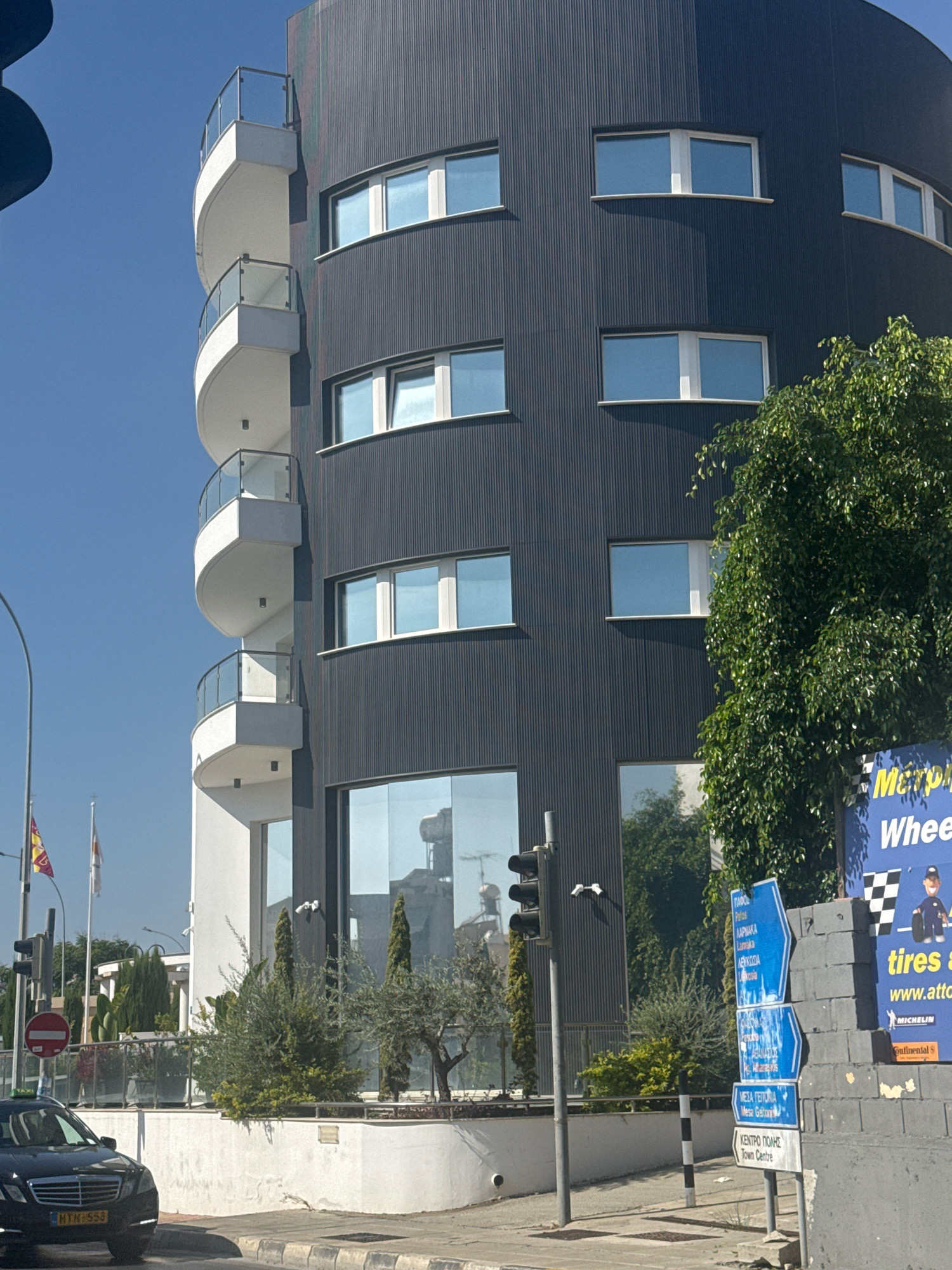 The image depicts a street view in Mesa Geitonia, Cyprus, dominated by a modern, cylindrical, dark-colored building with white, curved balconies. The building features numerous windows with light blue-tinted glass. In the foreground, a black car is parked on the right side of the street. To the right of the building, a tall green tree partially obscures a blue directional sign with white lettering in Greek and English, indicating various locations including "Mesa Geitonia" and "Town Centre." A black and white striped pole stands to the left of the sign. A traffic light with multiple lenses is visible on the left side of the image, along with two flags on poles. The sky is clear and blue.