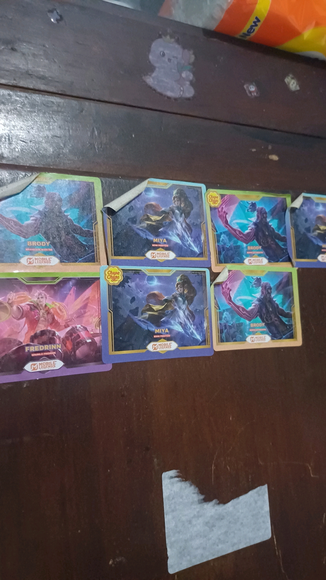 The image shows a collection of trading cards laid out on a dark wooden surface. The cards feature characters from the mobile game "Mobile Legends." Visible cards include "BRODY" with the subtitle "MARVELOUS MAESTRO" and "MIYA" with the subtitle "MOON PRIESTESS." Another card displays "FREDRINN" with the subtitle "SPARKLE FREDRINN." Some of the cards have "Chupa Chups" branding, indicating they are likely from a promotional campaign. The wooden surface appears to be a table or desk, showing some wear and possibly scratches. There are also some small stickers or adhesive remnants visible on the surface. The lighting suggests an indoor setting. The location is identified as Wonokerso, Indonesia.