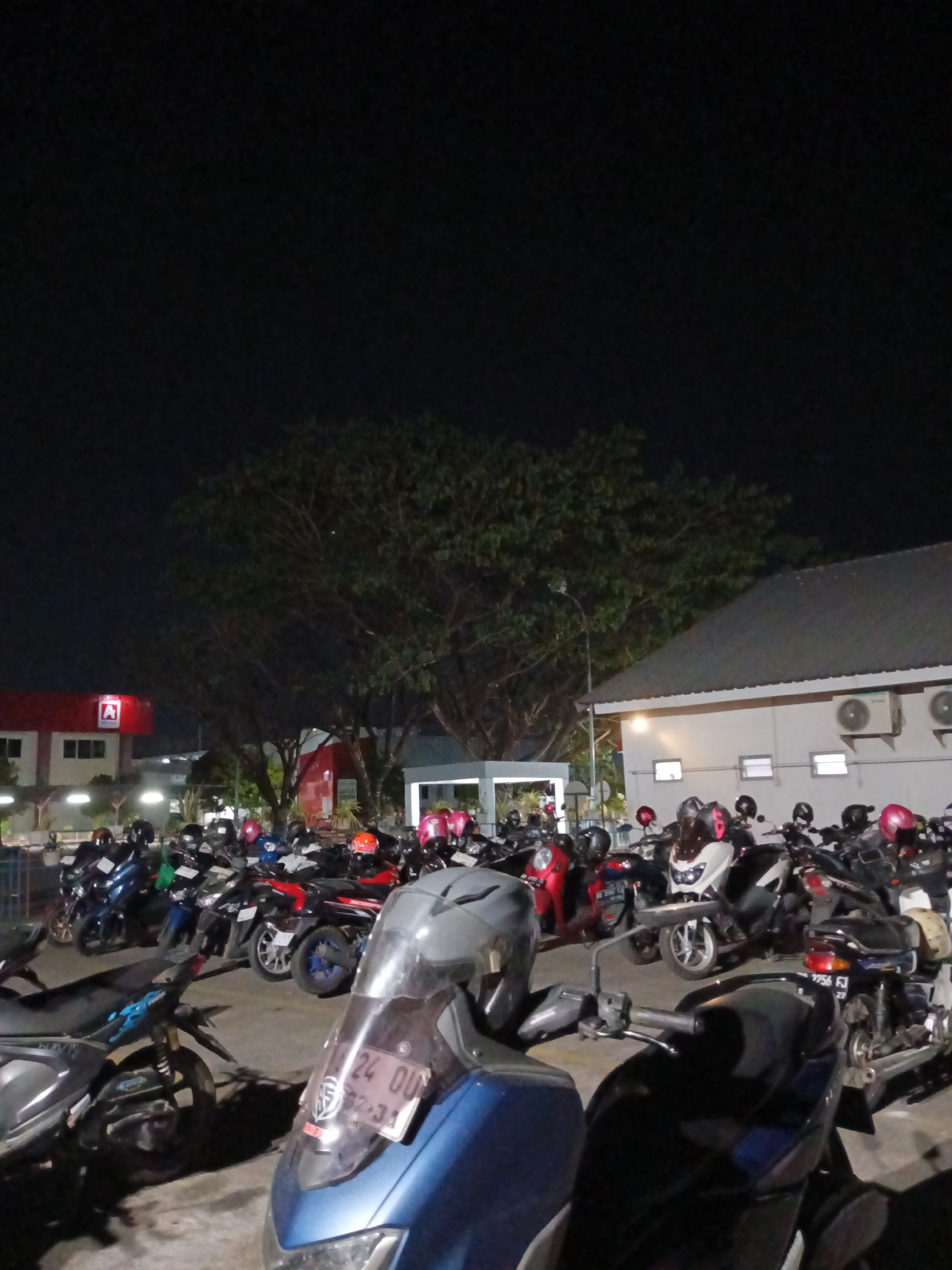 This image depicts a nighttime scene in a parking lot filled with motorcycles. In the background, there are buildings, including one with a red facade and a prominent red "A" logo, possibly indicating a business or gas station. Trees with dense foliage are visible against the dark night sky. Several motorcycles are parked closely together, some with helmets on their seats or handlebars. In the foreground, a blue scooter with a license plate reading "24 OU..." is prominently featured. The overall environment appears to be an urban setting in Makassar, Indonesia, likely a public area or commercial establishment's parking facility.