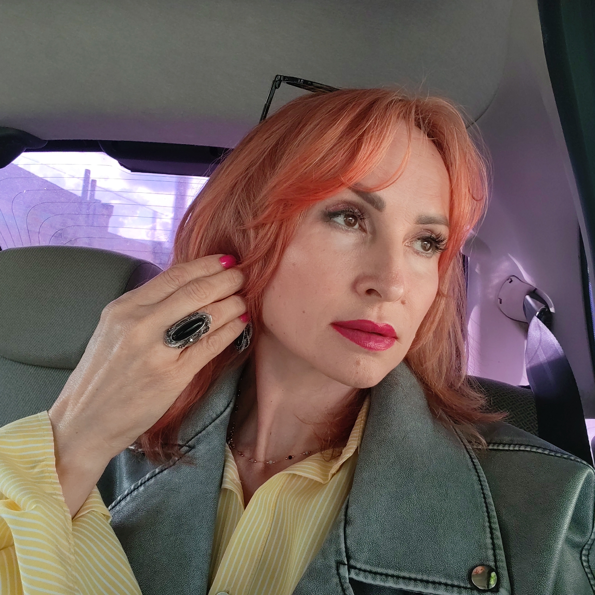 A woman with reddish-orange hair is sitting in the back seat of a car. She has fair skin, bright pink lipstick, and is wearing a large, oval black gemstone ring on her left ring finger. She's touching her hair with that hand. She is wearing a yellow and white striped shirt and a dark gray denim jacket. Her face is turned towards the right, looking out the window. The interior of the car is visible, including the back of the front seat, seatbelt, and a partially visible rear window. The location appears to be Chaikovsky, Russia, inferred from the distant buildings visible through the car's rear window.