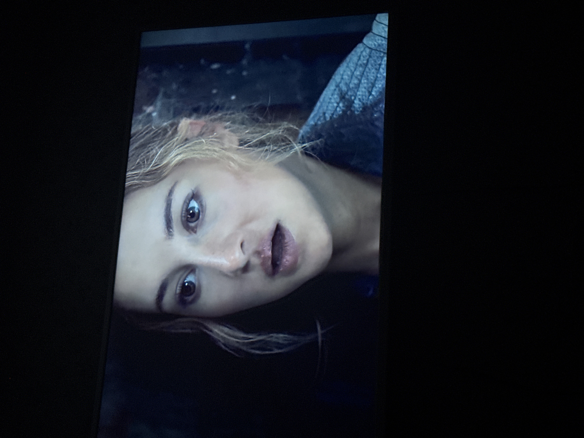 A close-up, angled shot depicts a woman with blonde hair and light skin appearing to look upward with an expression of surprise or shock. Her mouth is slightly open. She is wearing what appears to be a textured dark jacket. The background is dark and indistinct. The image seems to be a screen capture from a video or television. No specific location in Mytishchi, Russia, is discernible.