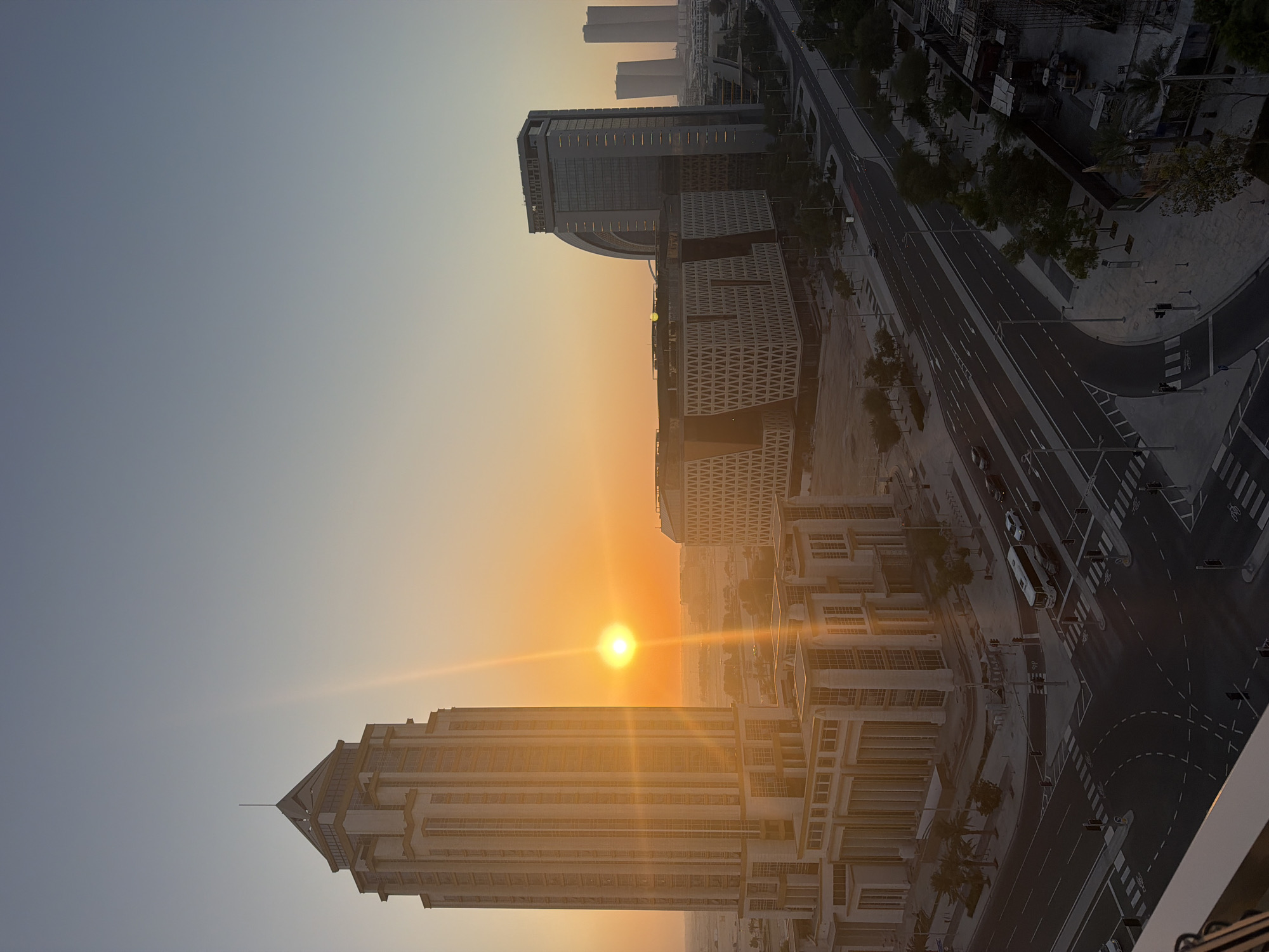 This image depicts a cityscape at sunset, captured from a high vantage point. The sun, a bright orb radiating golden light, hangs low in the sky, casting long shadows and illuminating the modern architecture. The scene is dominated by several large, contemporary buildings with geometric designs and numerous windows. Some buildings appear to be office complexes or residential towers. A multi-lane road with distinct markings is visible, featuring a few vehicles, including a bus. Sidewalks, streetlights, and pedestrian crossings are also present, suggesting an urban environment.
The time of day is clearly sunset, as indicated by the low position of the sun and the warm, orange and yellow hues of the sky. The weather appears clear, with no clouds visible. The overall atmosphere is serene and perhaps a little tranquil, typical of the transition from day to night in a developed urban area. The composition, with the buildings and road angled, gives a dynamic feel to the urban landscape. There is no visible text in the image.