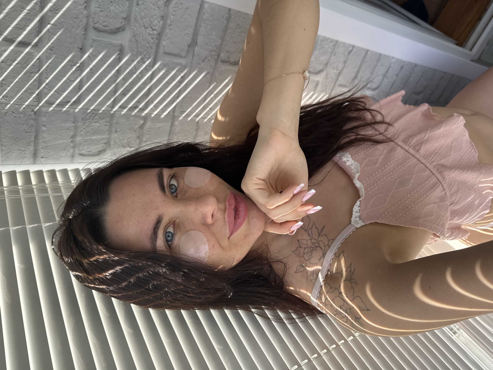 The image shows a woman with dark hair and light blue eyes, lying down with what appear to be eye masks under her eyes. She is wearing a light pink top with lace trim and has a floral tattoo on her collarbone. Her nails are manicured with a French tip style. Light from a window with blinds casts stripes across her face, hair, and body. The background includes a white brick wall and blinds. There is no specific information to determine if the photo was taken in the city of Chaikovsky, Russia.