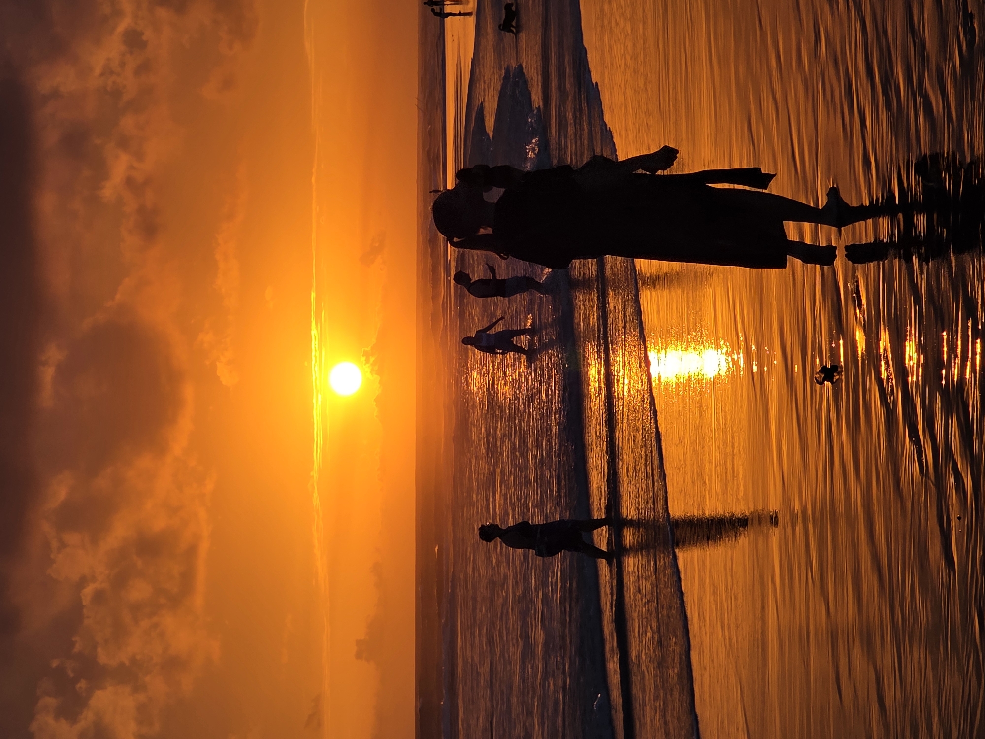 A group of people are silhouetted against a vibrant orange sunset on a beach in North Kuta, Indonesia. The sun is low on the horizon, casting a golden glow over the ocean and wet sand. Waves are gently rolling onto the shore, creating shimmering reflections. In the foreground, a person is walking towards the viewer, carrying what appears to be a child or a bag. Further out in the water, other figures are visible, some wading and others appearing to stand. One individual is looking at their phone. A dog is also present on the beach. The sky is filled with dramatic, textured clouds that are also illuminated by the setting sun. The overall scene conveys a sense of tranquility and natural beauty.
