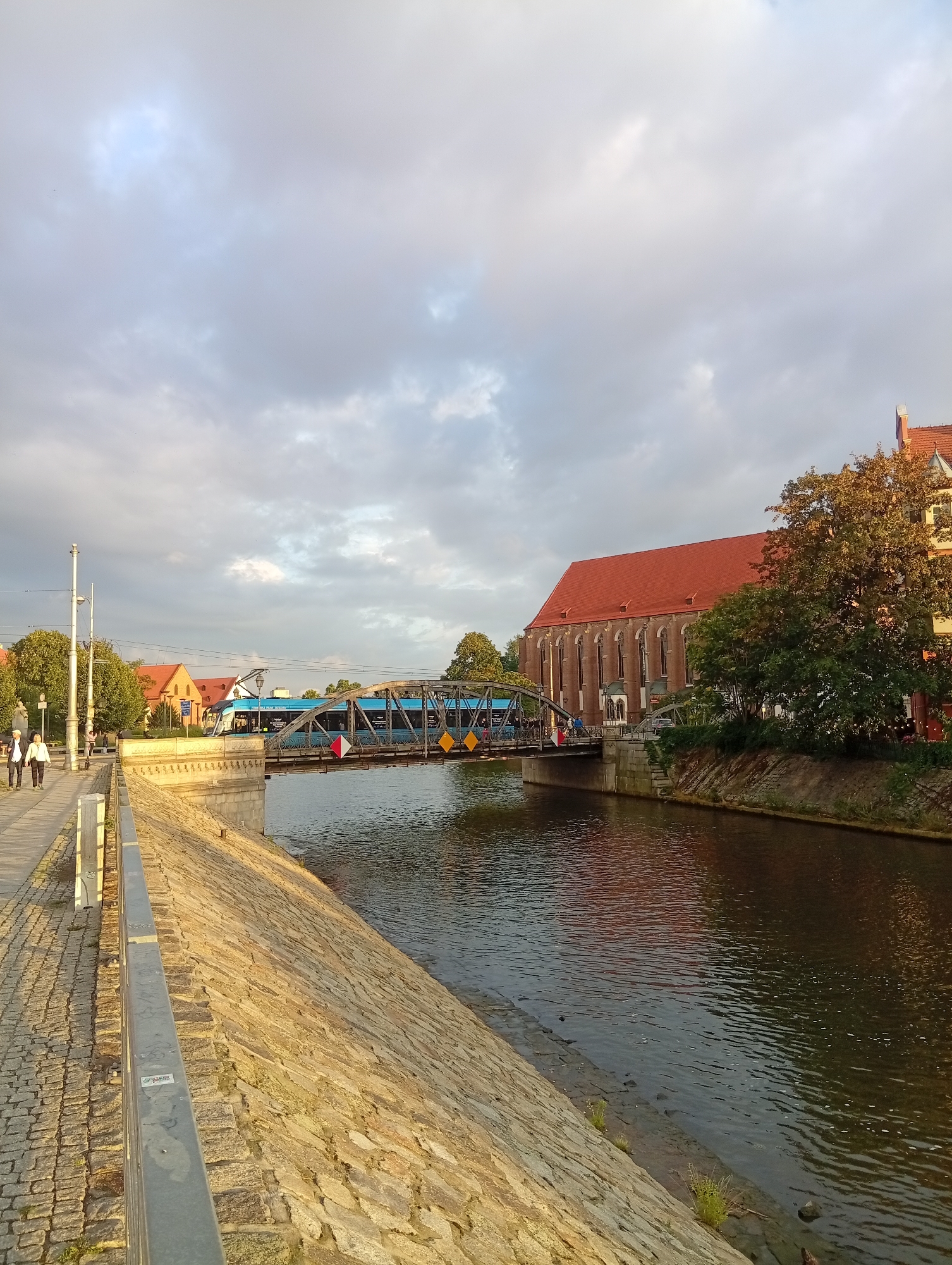 The image depicts a scene in Wrocław, Poland, along a riverfront promenade. A blue tram crosses a metal bridge spanning the river. On the left side of the image, a cobblestone walkway lines the river, bordered by a stone embankment. Two individuals are walking on the promenade. The buildings on the right feature red tiled roofs. The sky is cloudy.