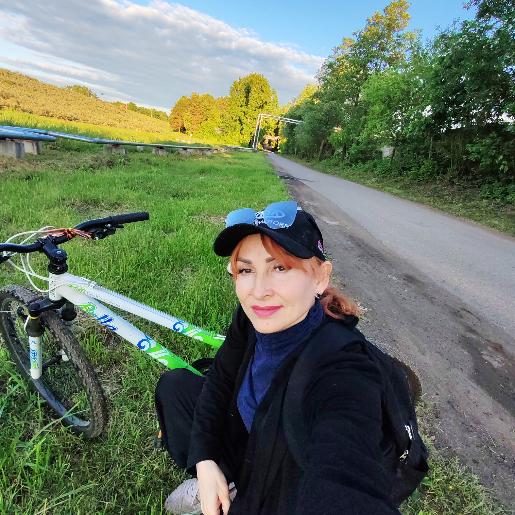 A woman poses outdoors with her bicycle in a rural or semi-rural setting near Chaikovsky, Russia. She has reddish-brown hair and is wearing a black baseball cap with blue mirrored sunglasses resting on top, a black jacket over a dark blue turtleneck, and light-colored sneakers. She is crouching on a grassy verge next to a narrow paved road, looking directly at the camera with a gentle smile.
Her bicycle, a white mountain bike, is next to her in the grass. Visible text on the bike frame includes "Bella" and "CROSS COUNTRY." The word "MOTORS" is visible on her cap.
The background features a gently sloping, grassy hill on the left with a series of large, grey pipes running along its base. Beyond the pipes, the hillside is covered in vegetation and trees. To the right of the road, a dense line of green trees extends into the distance. The sky above is clear and blue with scattered white clouds. The warm, golden light suggests late afternoon or early evening, indicating the "golden hour," with some trees in the background illuminated by the setting sun. The scene conveys a peaceful moment, likely during a leisurely bike ride.