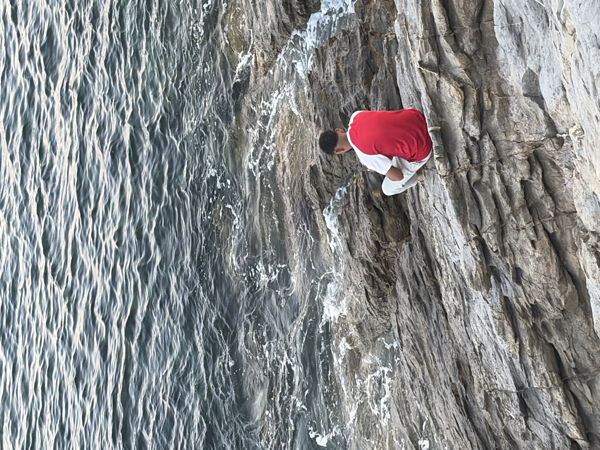 The image captures a person sitting on rocks by the water's edge, likely in Imperia, Italy, given the context. The person, seen from behind, is wearing a red and white t-shirt and light-colored pants. They are seated on layered grey rocks that slope towards the choppy, dark blue water. Small white waves are breaking against the rocks, creating foam and white streaks in the water. The texture of the rocks is rough and stratified. The scene is natural and appears to be an outdoor coastal environment.