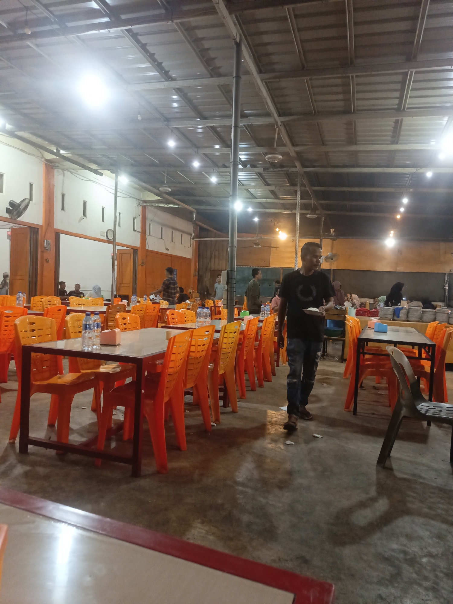 The image depicts the interior of a large, open-air restaurant or eatery located in Lhokseumawe, Indonesia. The space is covered by a corrugated metal roof supported by exposed beams and metal framing. Numerous fluorescent lights are suspended from the ceiling, illuminating the scene.
The restaurant is furnished with many tables and chairs. The tables appear to be dark-colored with metal frames, and most are surrounded by bright orange plastic chairs. Some tables have water bottles and small containers placed on them.
Several individuals are visible within the restaurant. In the foreground, a man wearing a black t-shirt with a circular graphic and blue jeans walks towards the right, holding a plate of food. In the background, to the left, a group of people are seated at a table, including a person wearing a headscarf. Further back, other patrons and staff can be seen.
The floor is concrete and shows some wear. The walls are painted white with orange accents. The overall atmosphere suggests a busy, casual dining establishment.