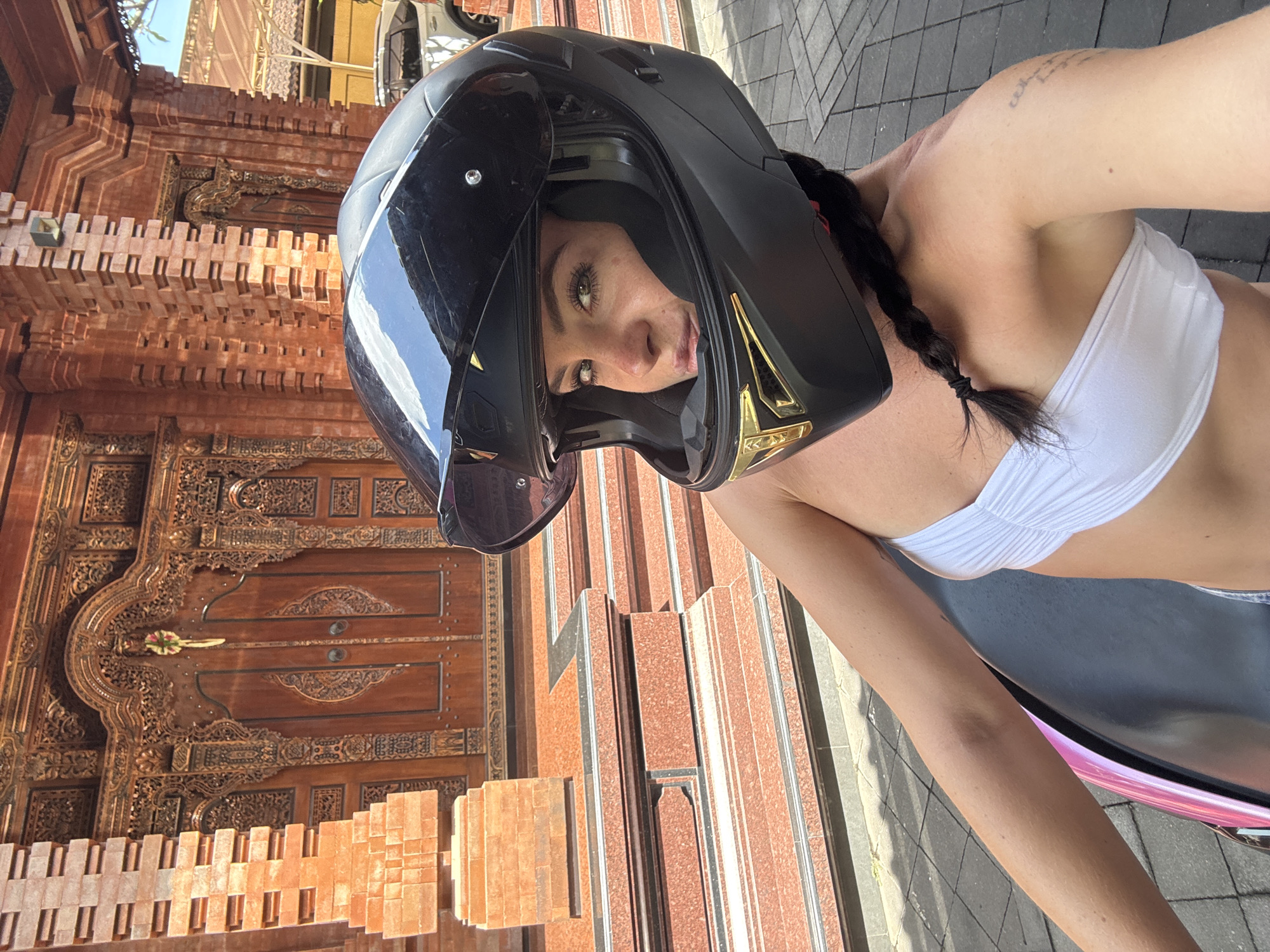 A woman wearing a black helmet and a white bikini top poses for a selfie in front of a building with ornate wooden doors and intricately carved details. The building appears to be made of red brick and stone. The woman has her hair in a braid, and her eyes are visible through the helmet's visor. She is sitting on what looks like a motorcycle, indicated by the handlebars and part of the seat visible in the foreground. The scene suggests it is daytime, with sunlight casting shadows on the ground. The location is identified as North Kuta, Indonesia, which aligns with the architectural style of the building. There is no visible text in the image.